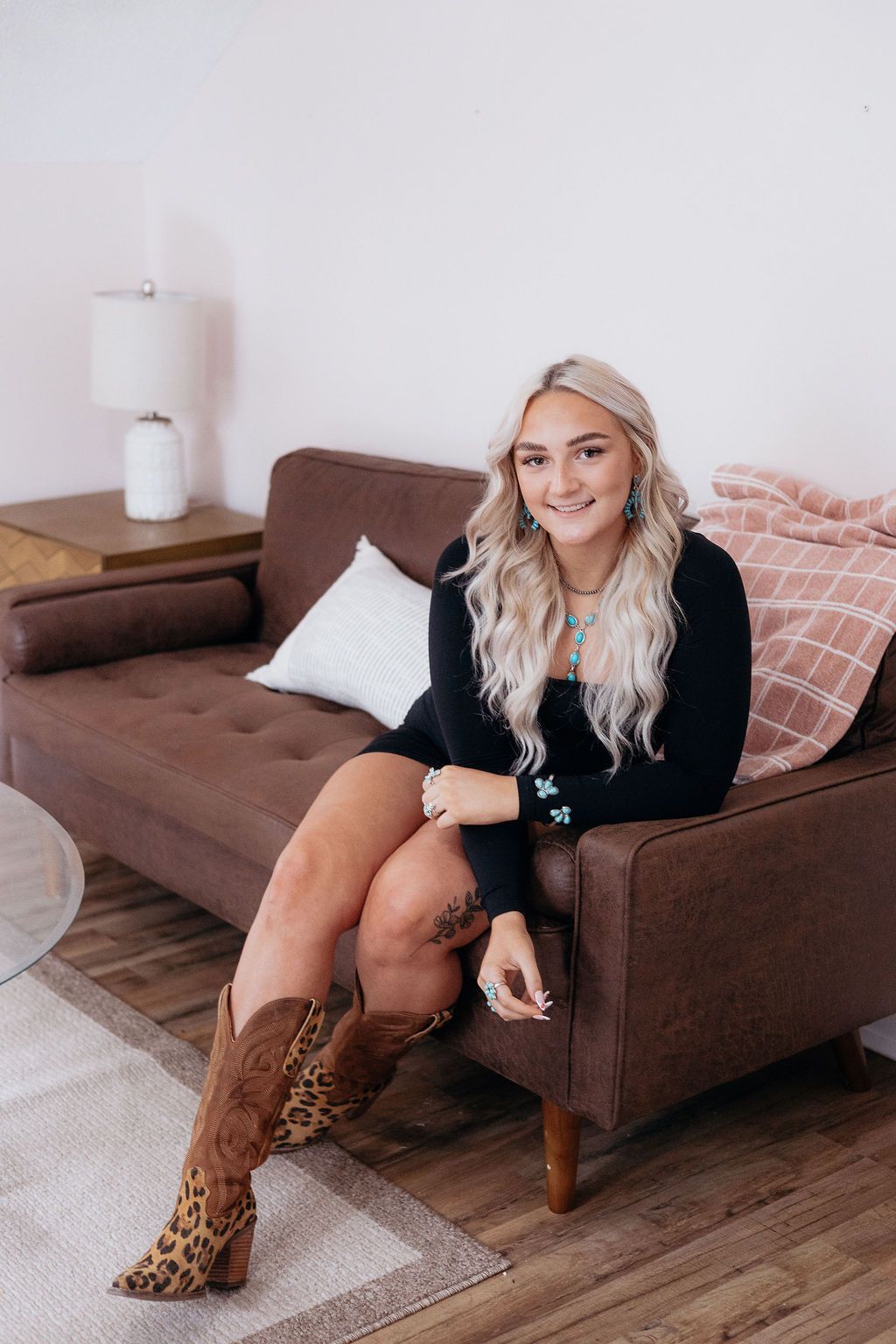 Woman with long blonde hair, smiling, seated on brown couch wearing leopard print boots, black sweater.