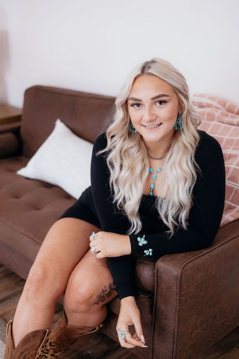 Blonde woman smiling on a brown couch, wearing a black dress, turquoise jewelry, and cowboy boots.