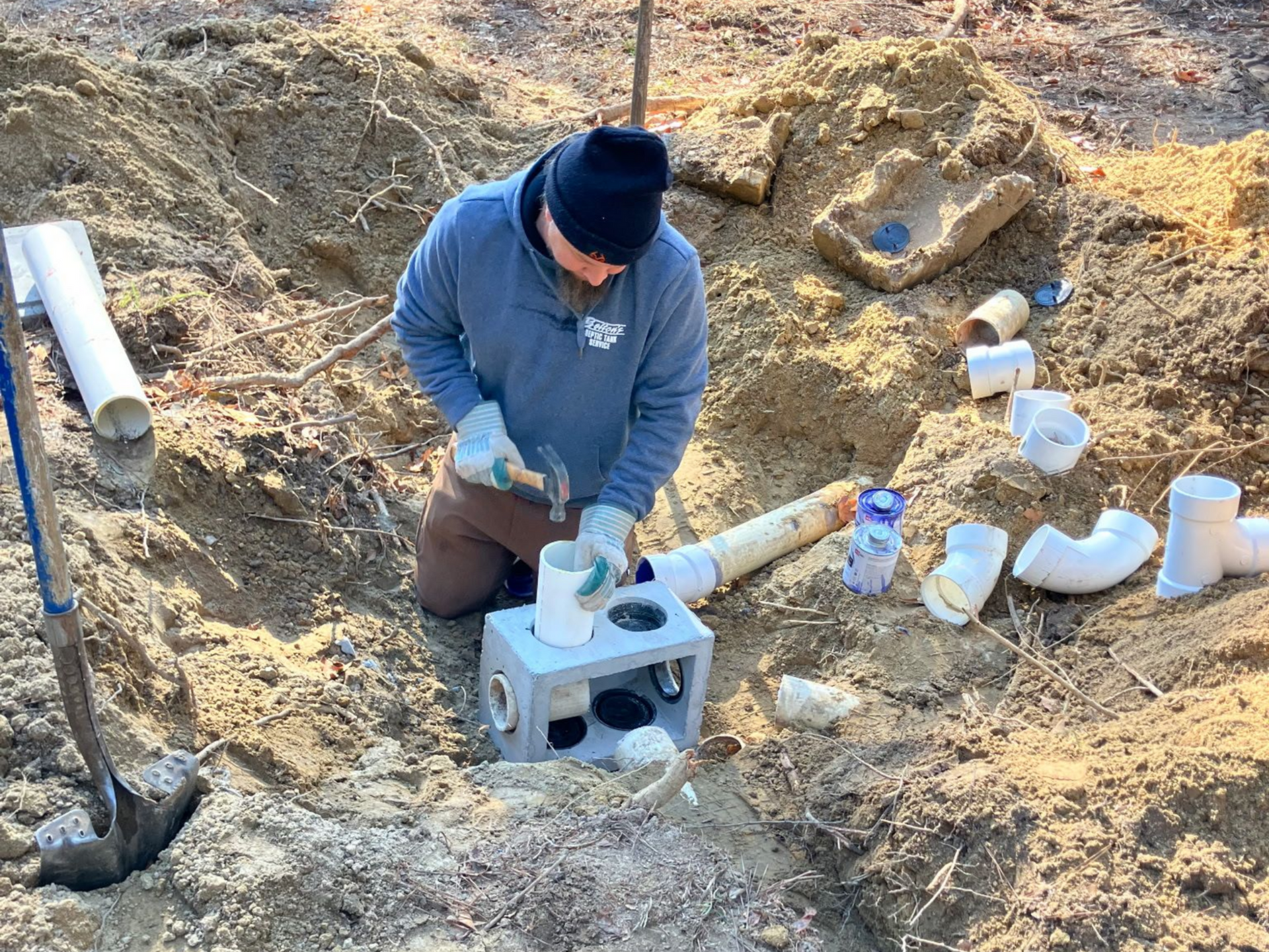 Man kneeling in a dirt pit, hammering plumbing fittings, surrounded by pipes and tools.
