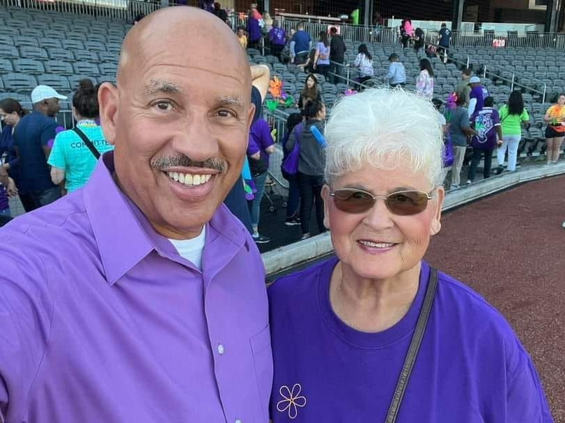 Two people in purple shirts smile, posing for a selfie at a stadium.