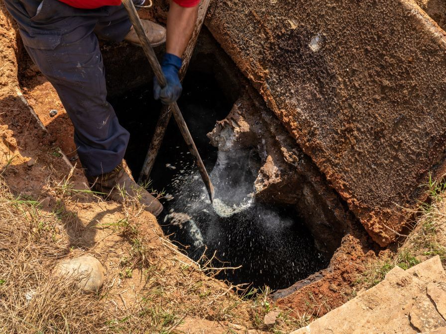 Person in blue gloves and pants cleaning a dark, open septic tank; brown soil and concrete frame.