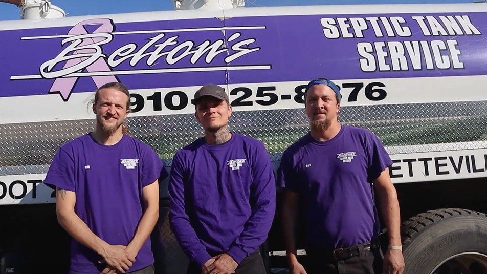 Three men in purple shirts stand in front of a septic tank truck with the company name, Belton's, and phone number.