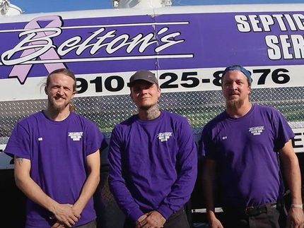 Three men in purple shirts are standing in front of a septic service truck