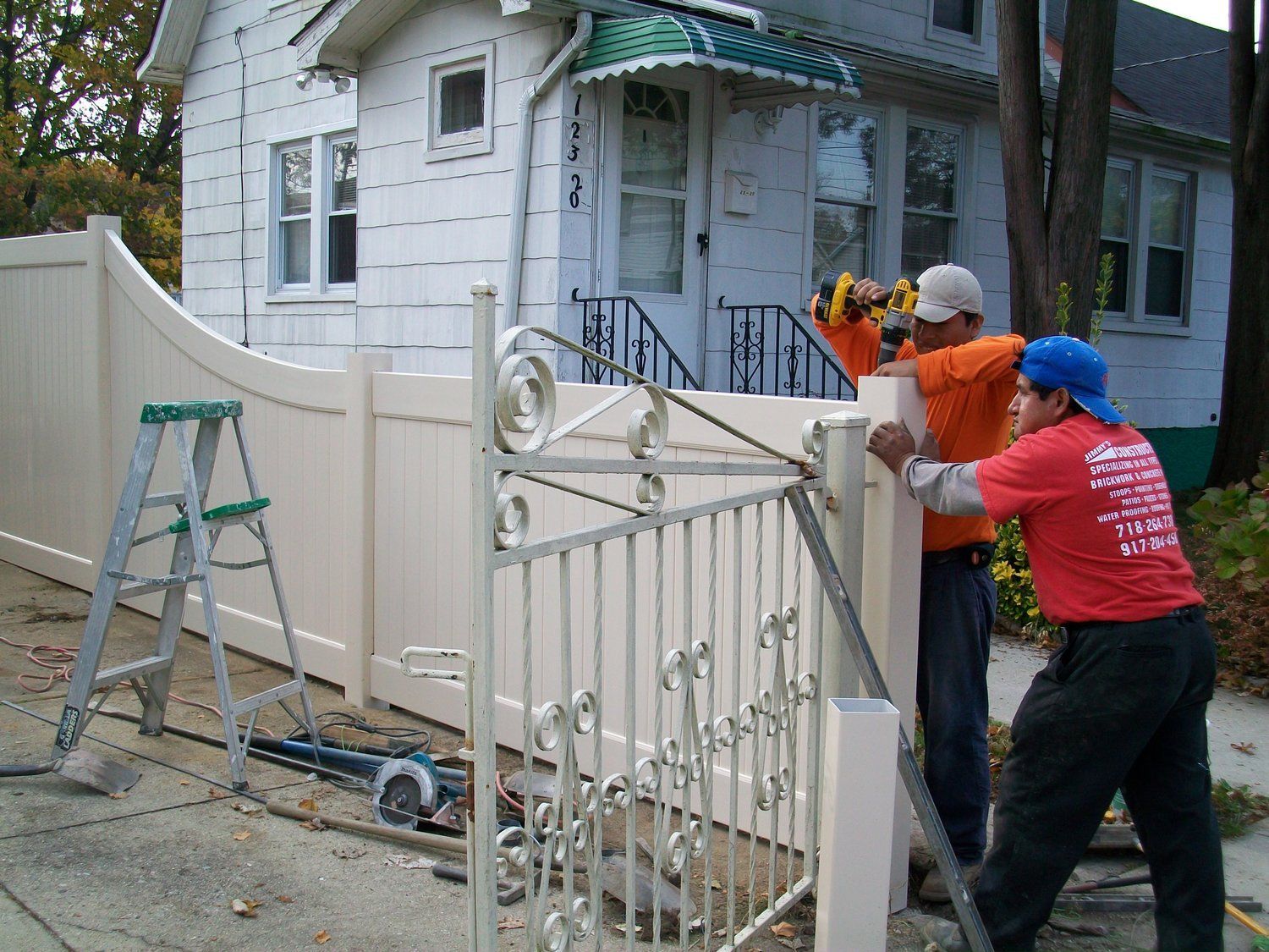 Two men are working on a white fence in front of a house