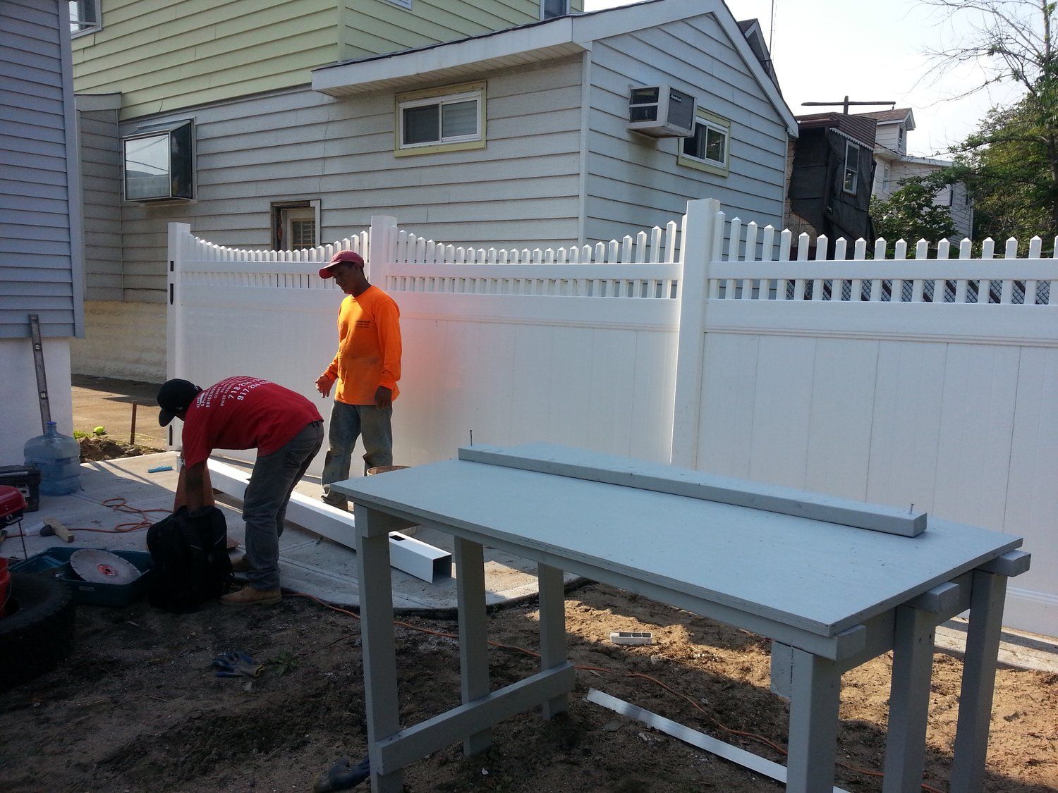 Two men are working on a white fence in front of a house