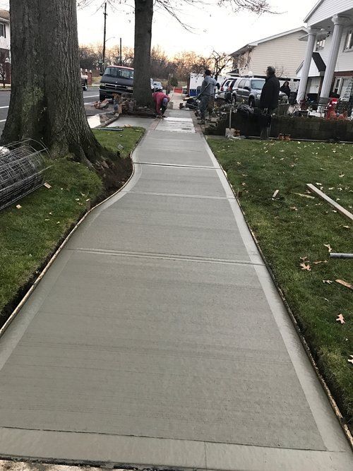 A concrete sidewalk leading to a house with cars parked on the side of the road.