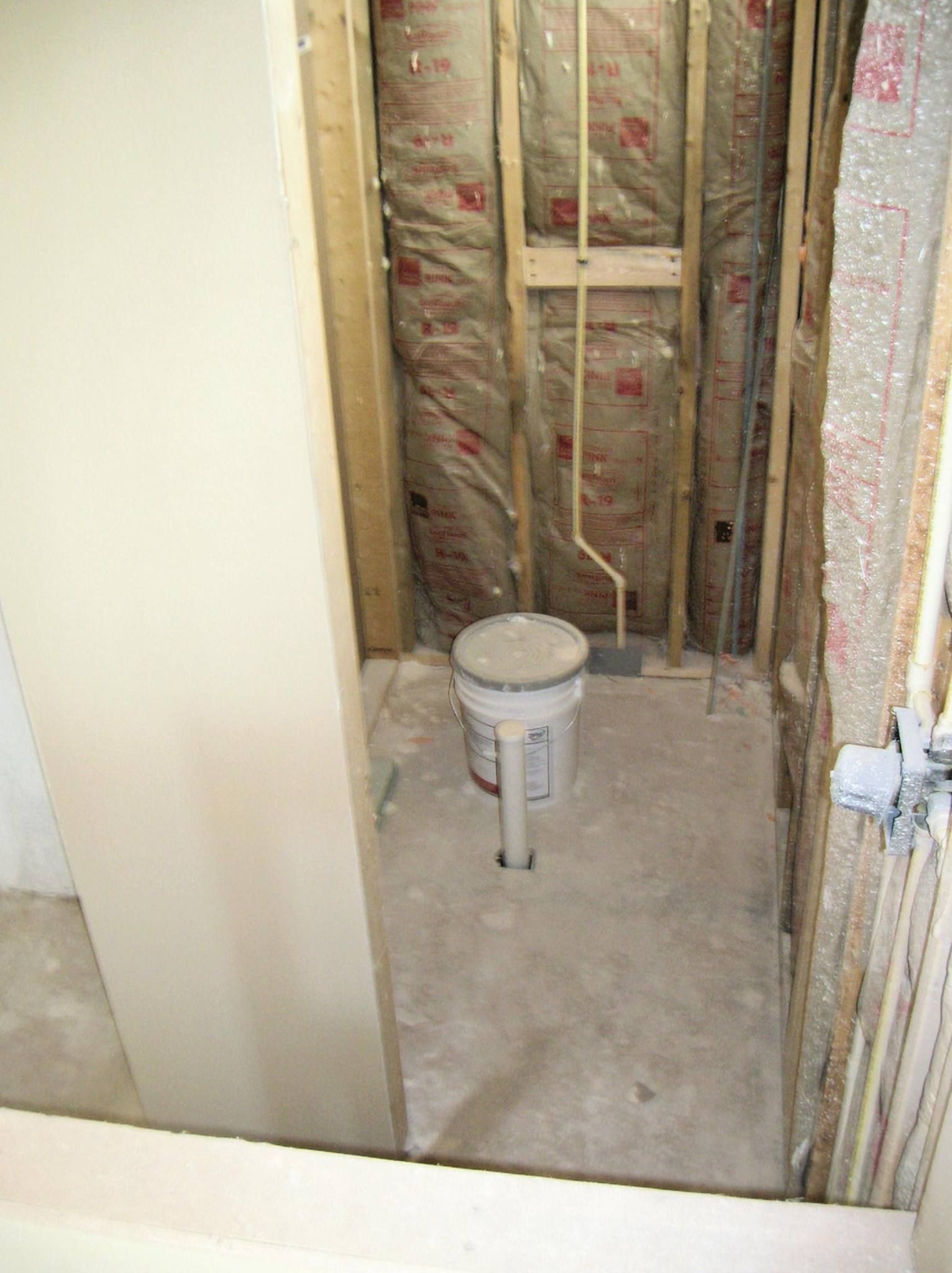 Bathroom renovation in progress, framing, insulation, concrete floor, pipe, and bucket visible.