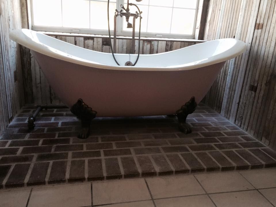 Pink claw-foot bathtub on a brick floor, set against wood-paneled walls, with a window and shower fixtures.
