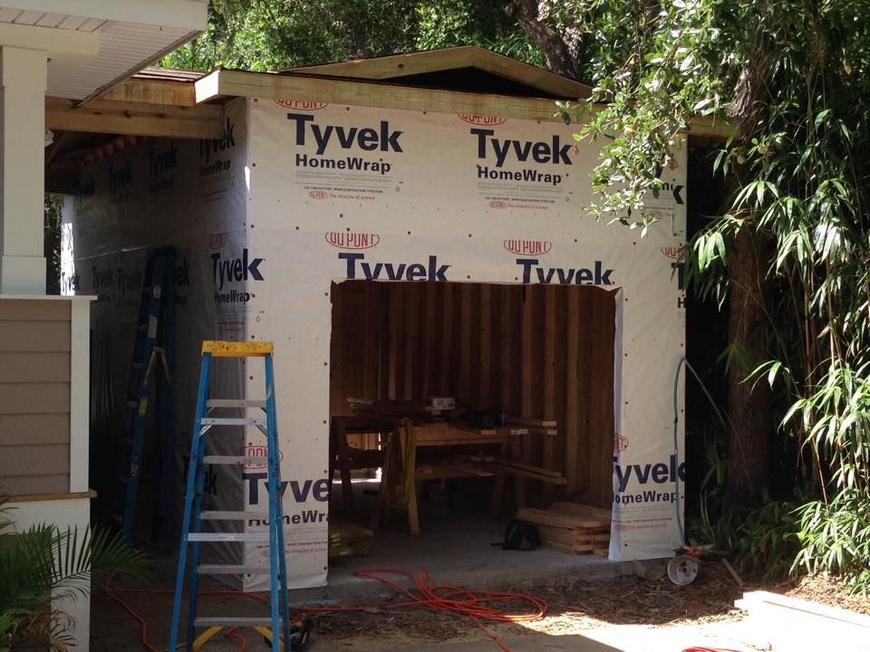 Construction site: Shed frame wrapped in white Tyvek, partially constructed, with ladder and workbench inside.