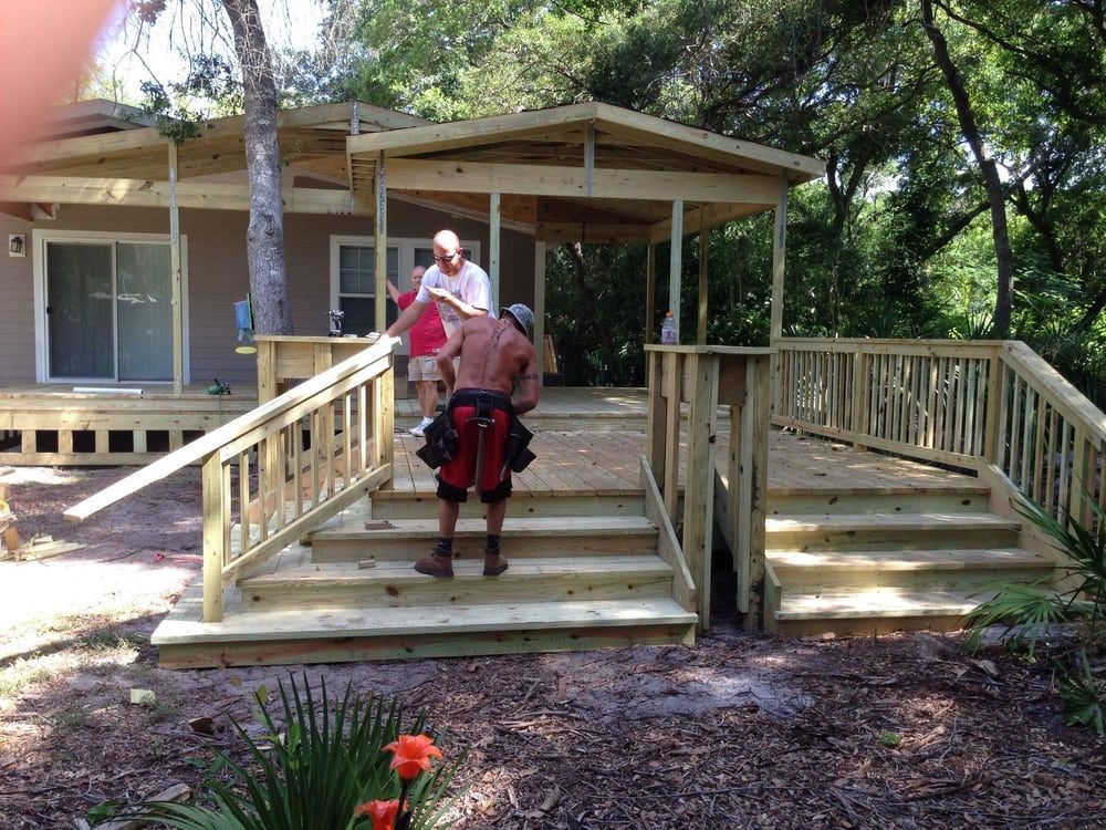 Men building a wooden deck with stairs and a small roofed section. Trees surround the house.