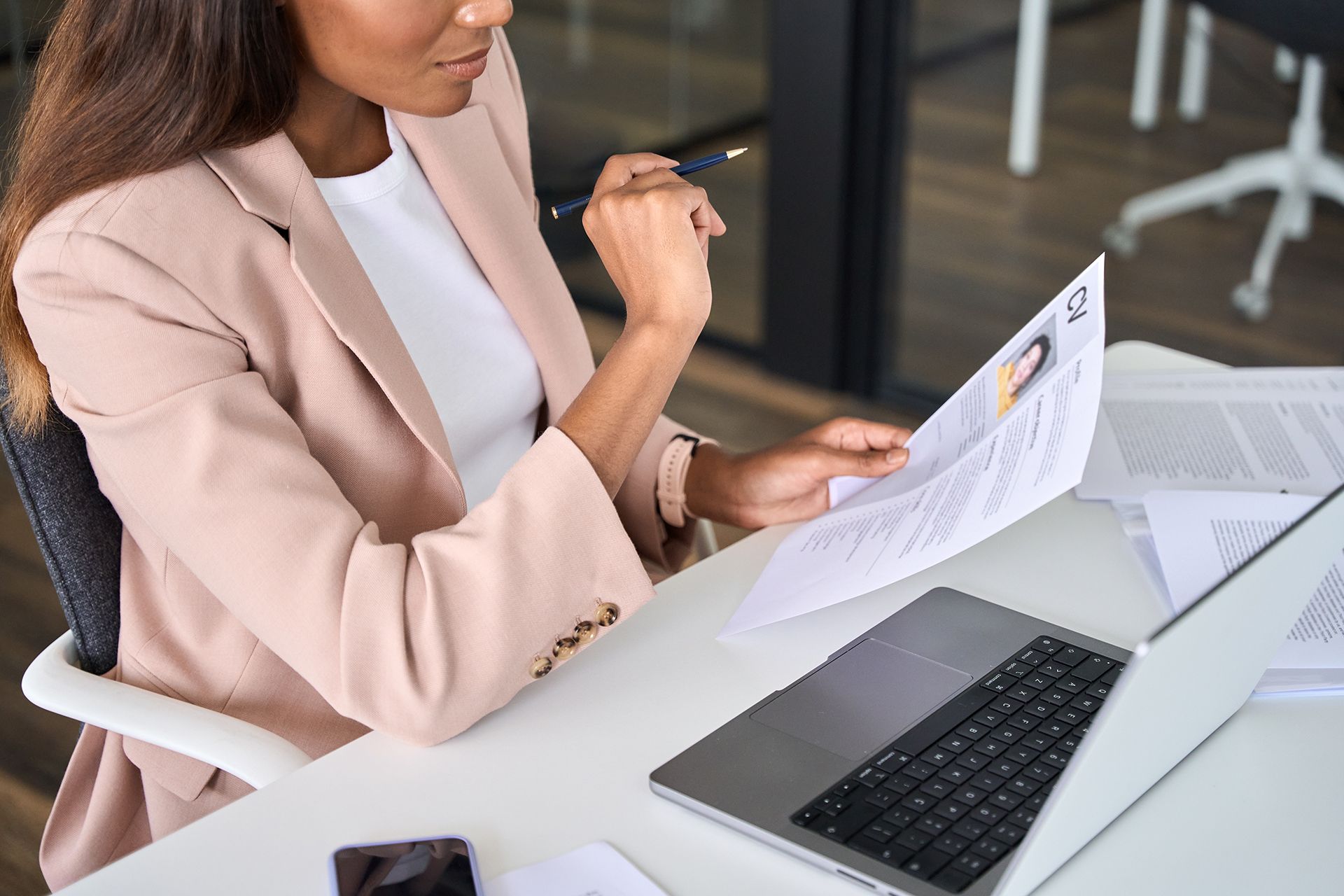 A woman is sitting at a desk with a laptop and looking at a resume.