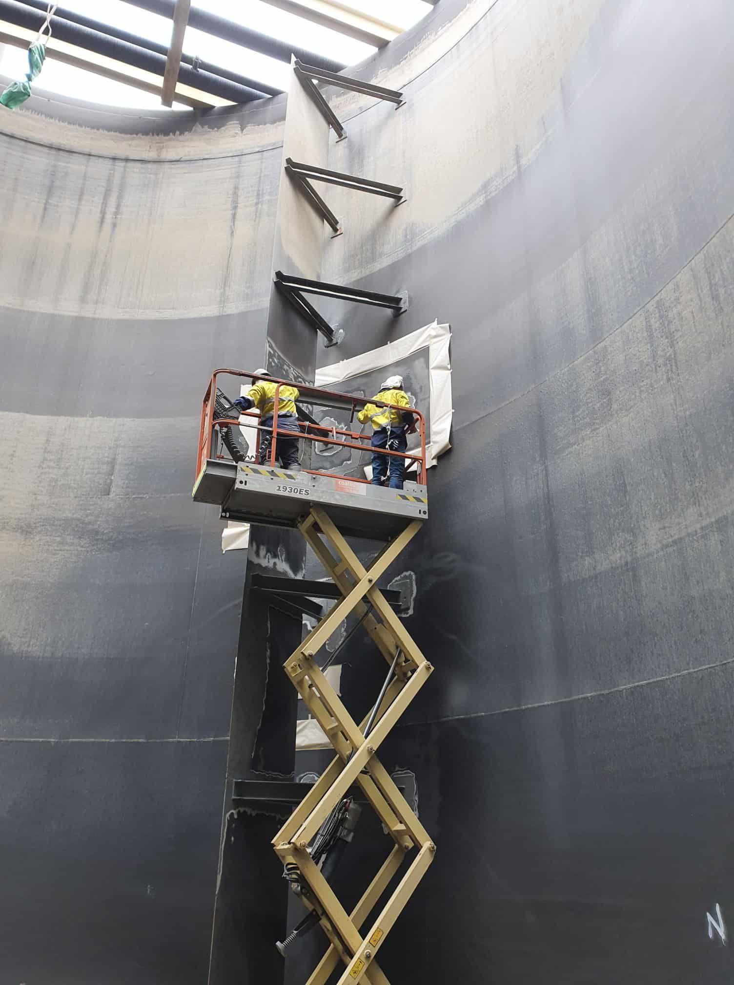 Two Men Are Standing on a Scissor Lift in a Large Room — Waters Abrasive Blasting Services In Shaw, QLD