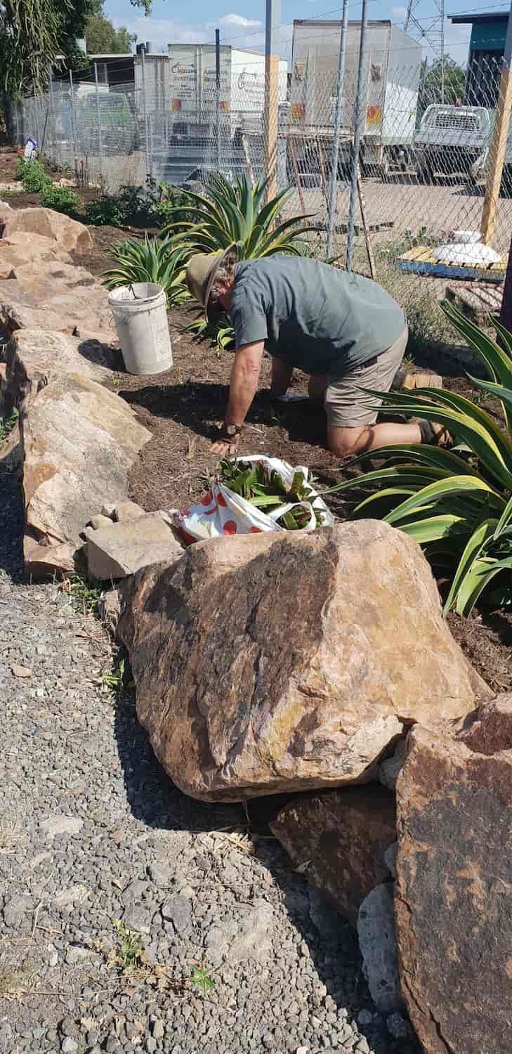 A Man is Kneeling Down in the Dirt Next to a Large Rock — Waters Abrasive Blasting Services In Whitsundays, QLD