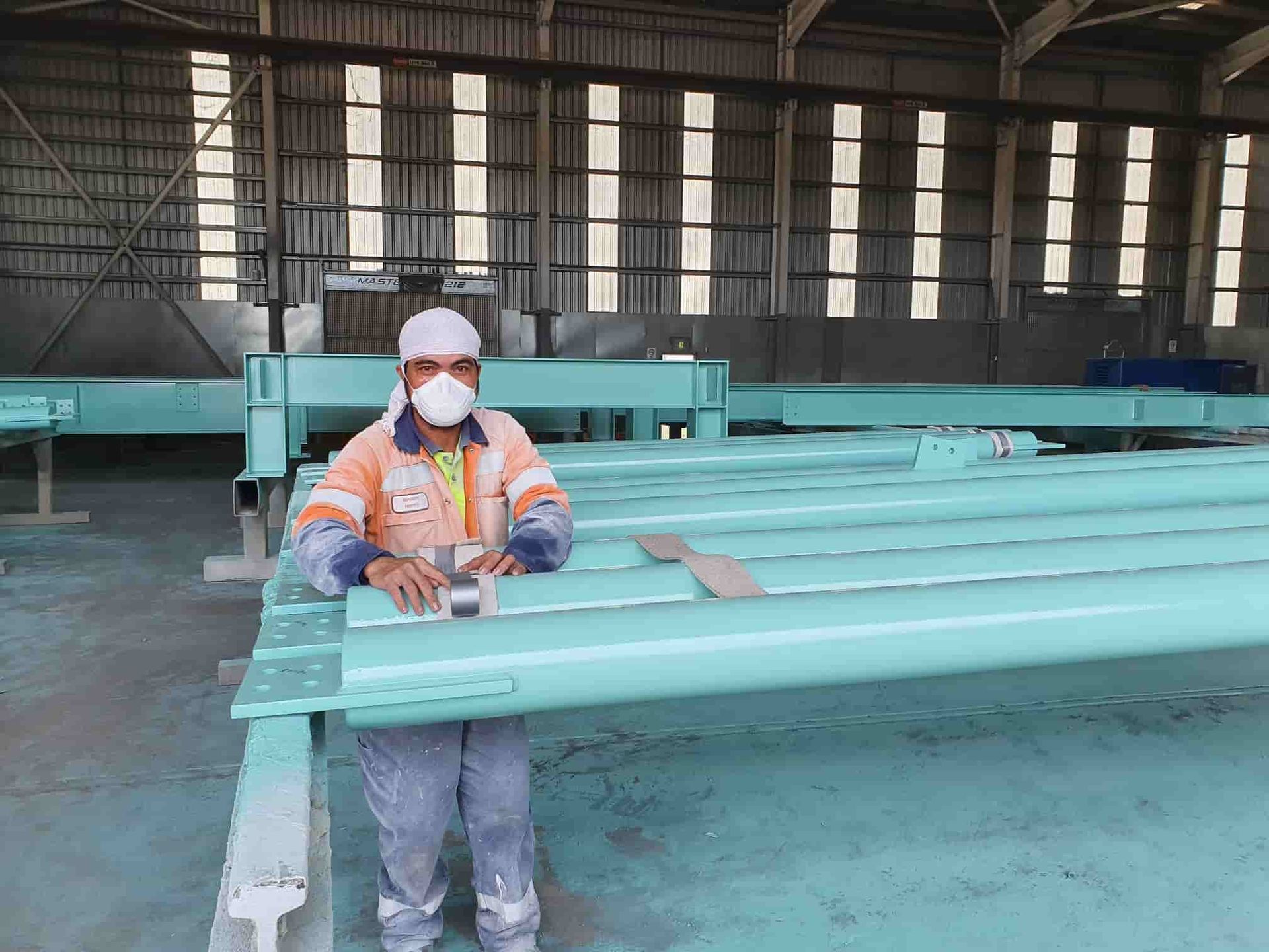 A Man Wearing a Mask is Standing Next to a Row of Green Pipes in a Factory — Waters Abrasive Blasting Services In Shaw, QLD