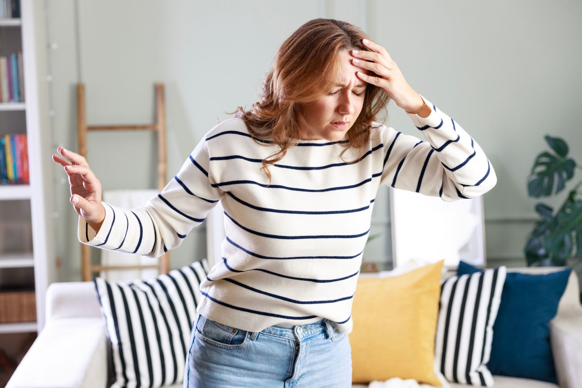 A woman in a striped sweater is standing in front of a couch holding her head.