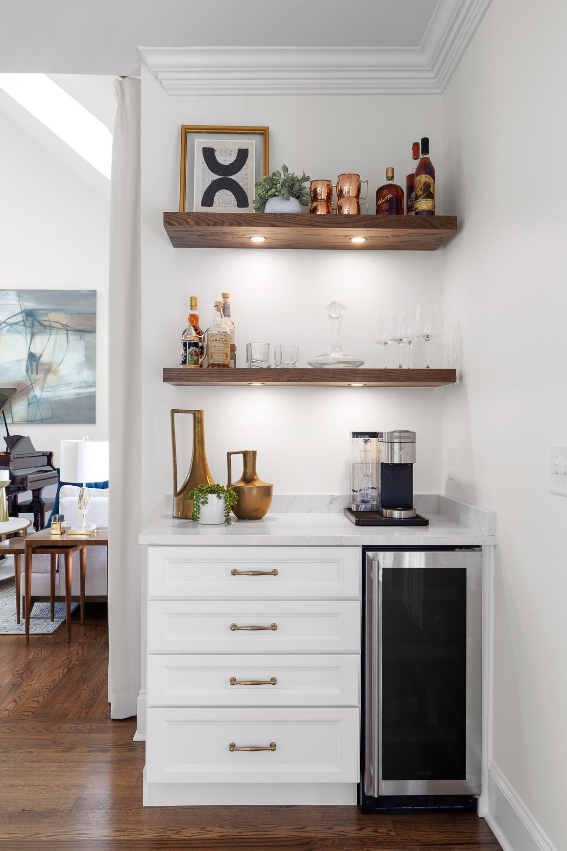 Small white home bar with liquor, coffee machine, shelves, and mini-fridge; dark wood floors and white walls.
