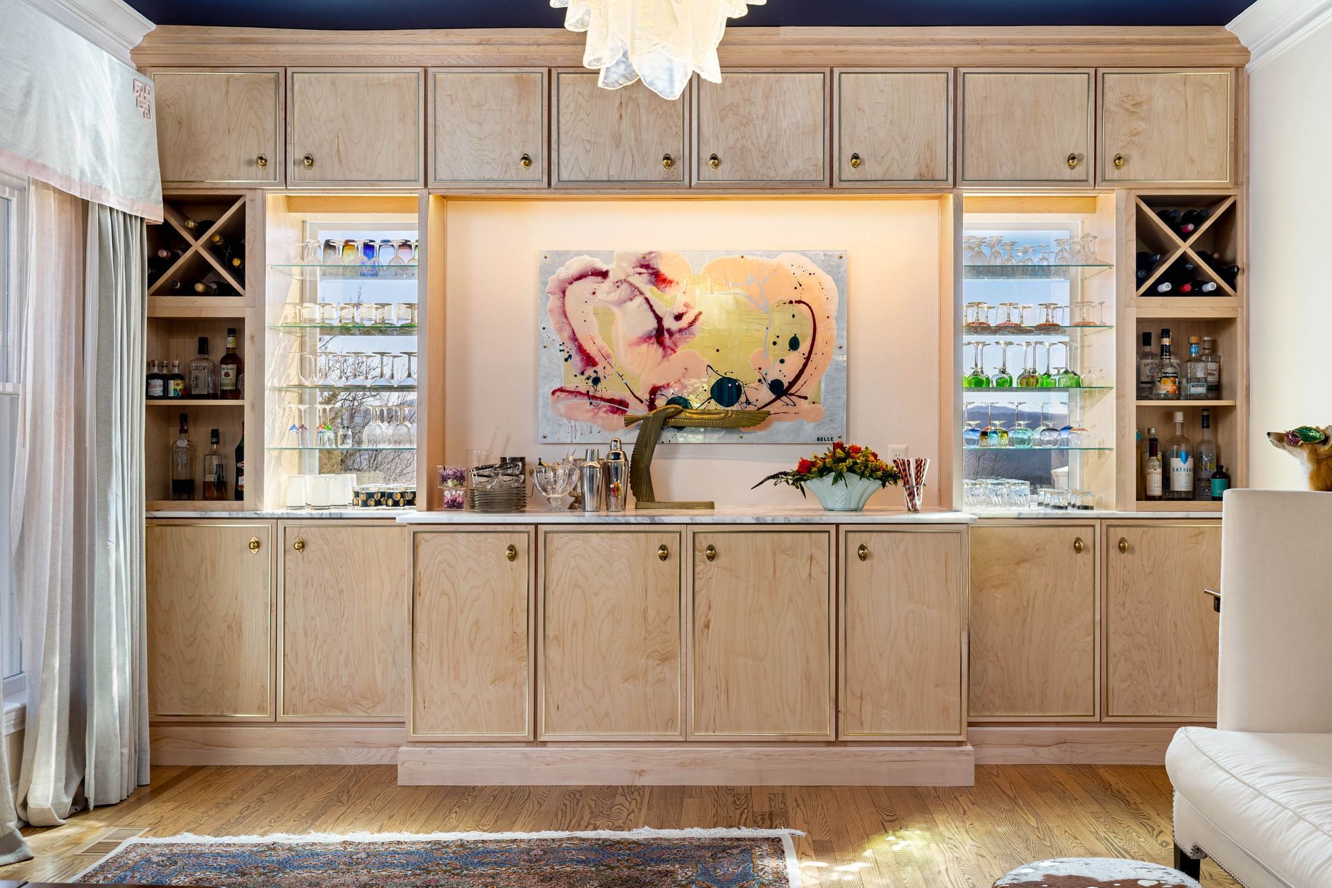 A built-in bar area with light wood cabinets, artwork, and shelving with glassware and bottles.