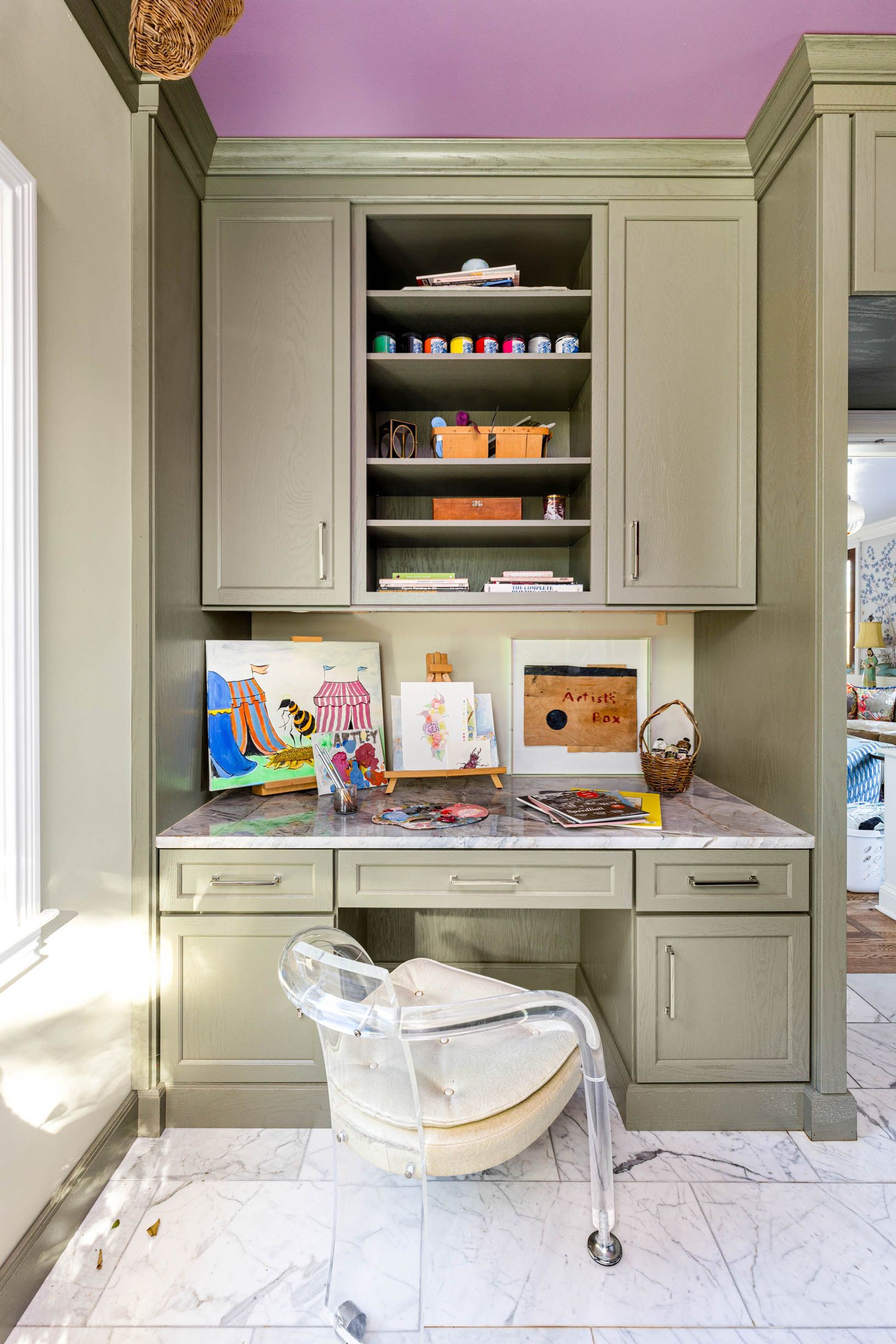 A home art studio nook with green cabinets, desk, a clear chair, and art supplies on a white marble floor.