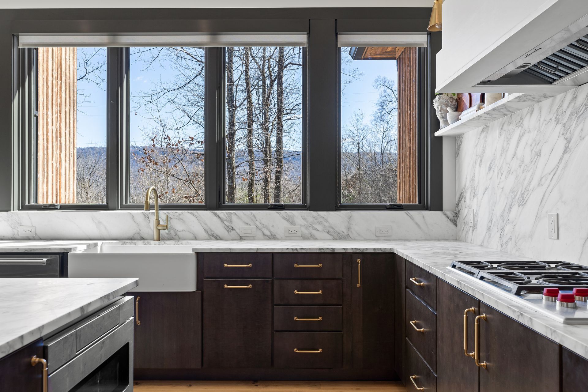 Modern kitchen with dark cabinets, marble counters, and a view of trees through the window.