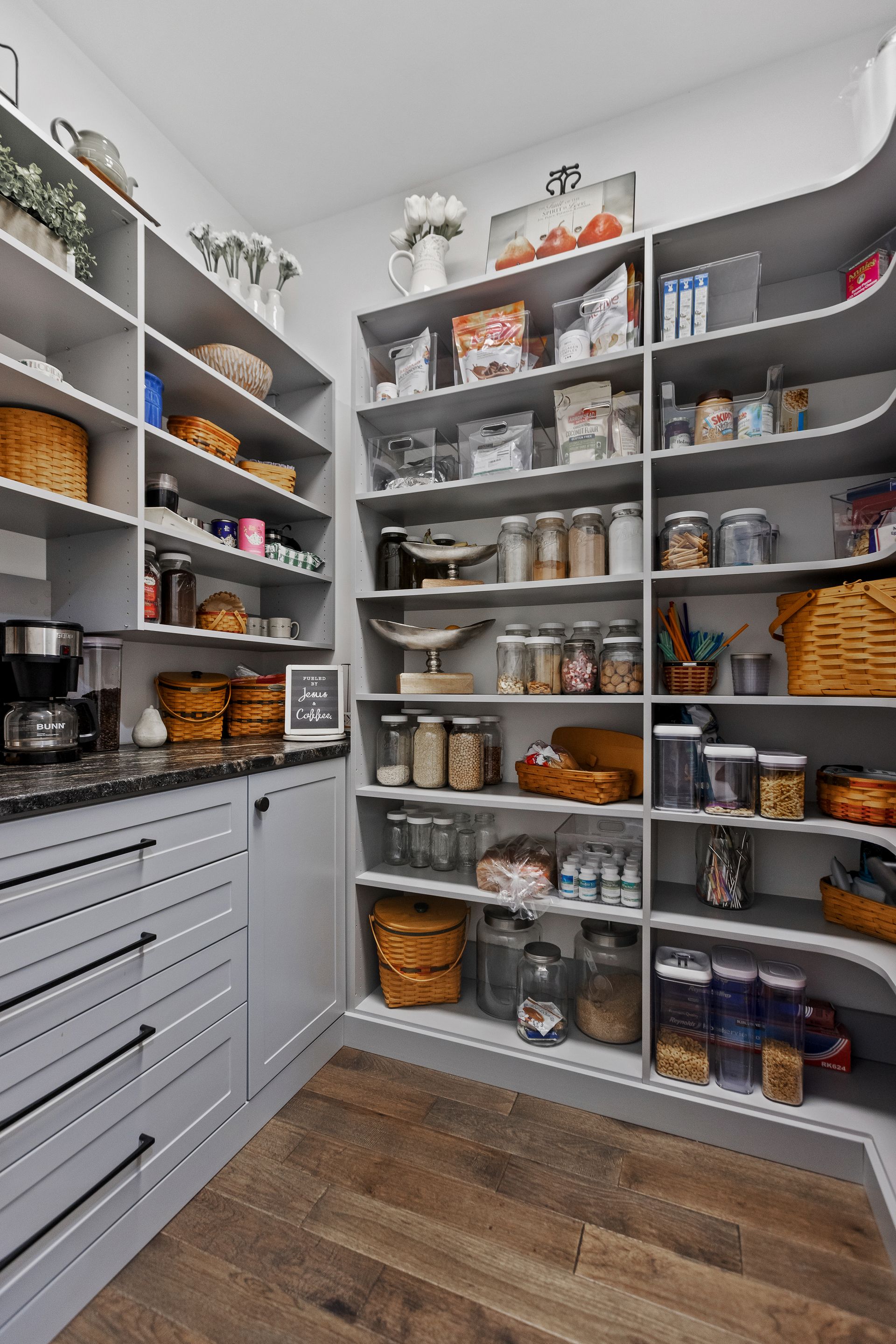 A pantry with gray shelves filled with food items, baskets, and jars, against a hardwood floor.