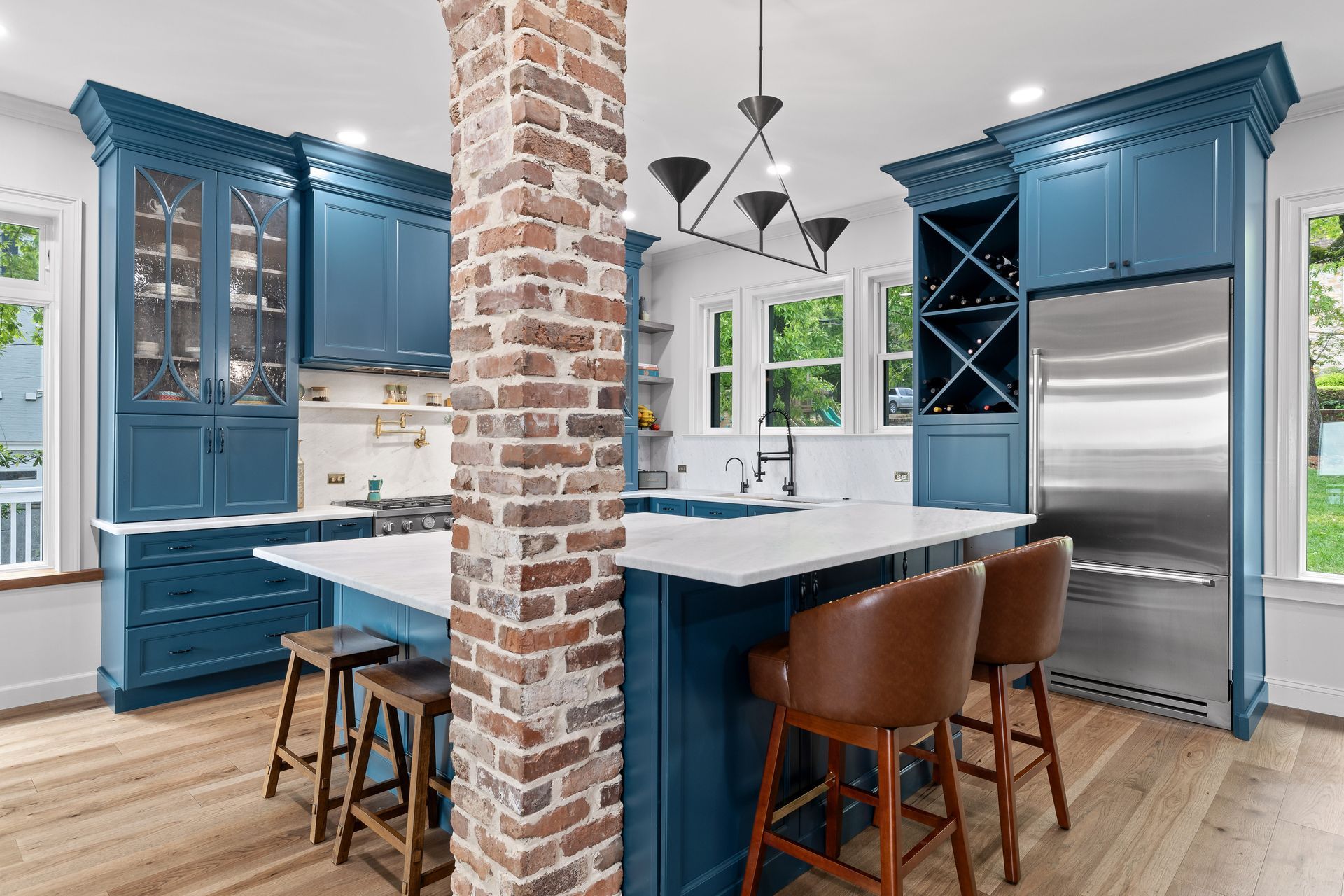 Blue kitchen with brick pillar, white island, wooden floors, and brown leather barstools.
