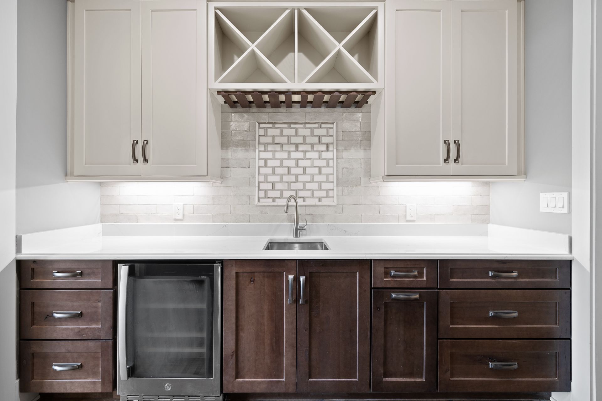 A home bar with light upper cabinets, wine rack, and dark lower cabinets with a stainless steel beverage cooler.