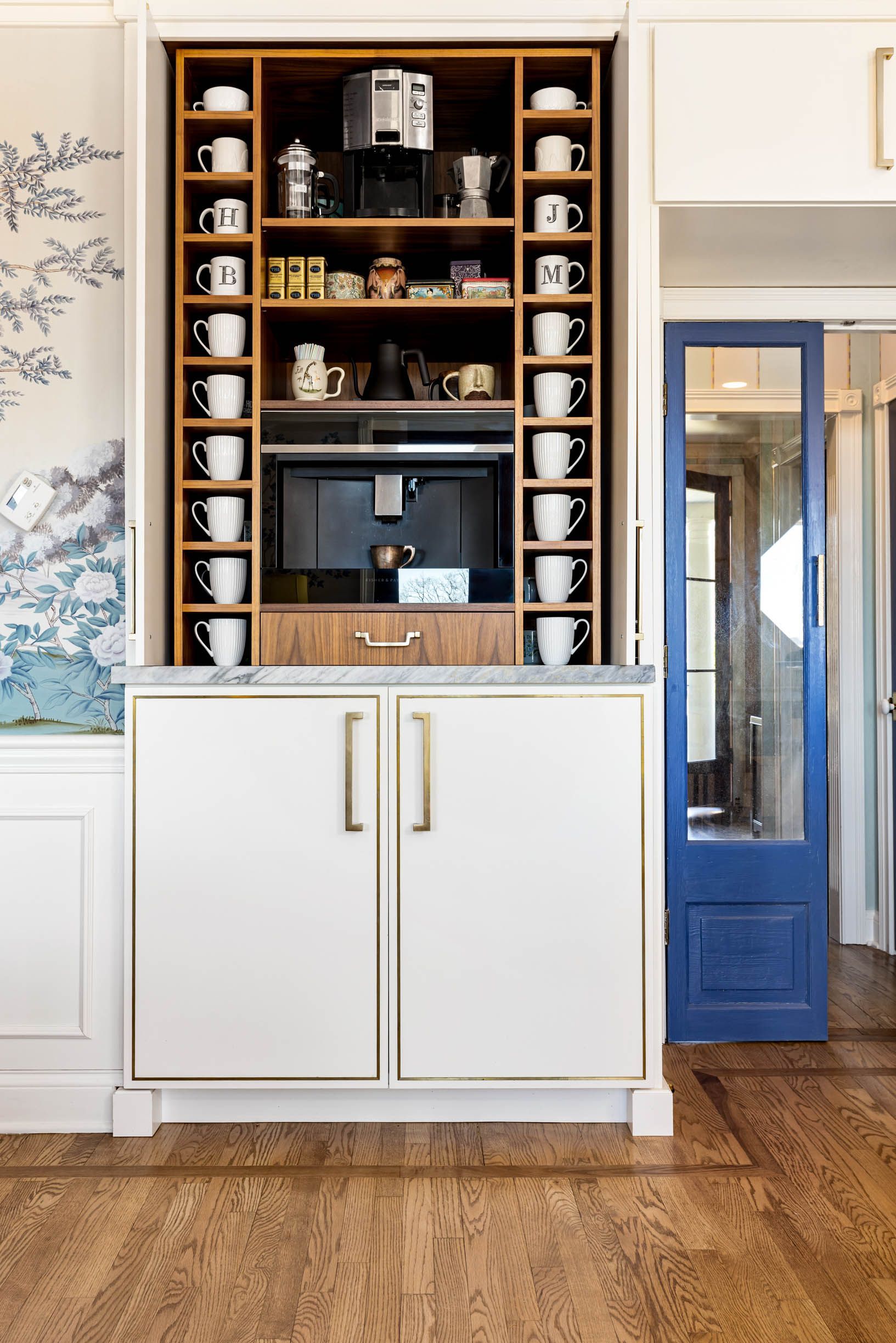 Built-in coffee bar with white cabinets, wood shelving, coffee mugs, and a blue door nearby.