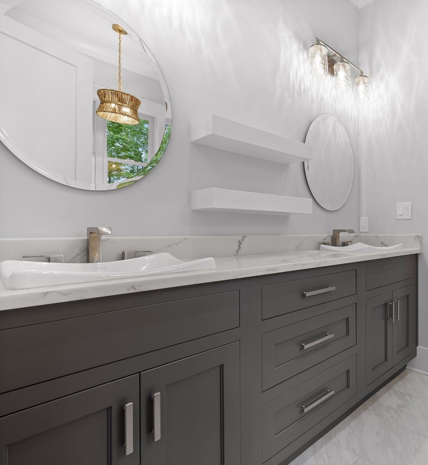 Bathroom with dark gray cabinets, white countertops, round mirrors, and overhead lighting.