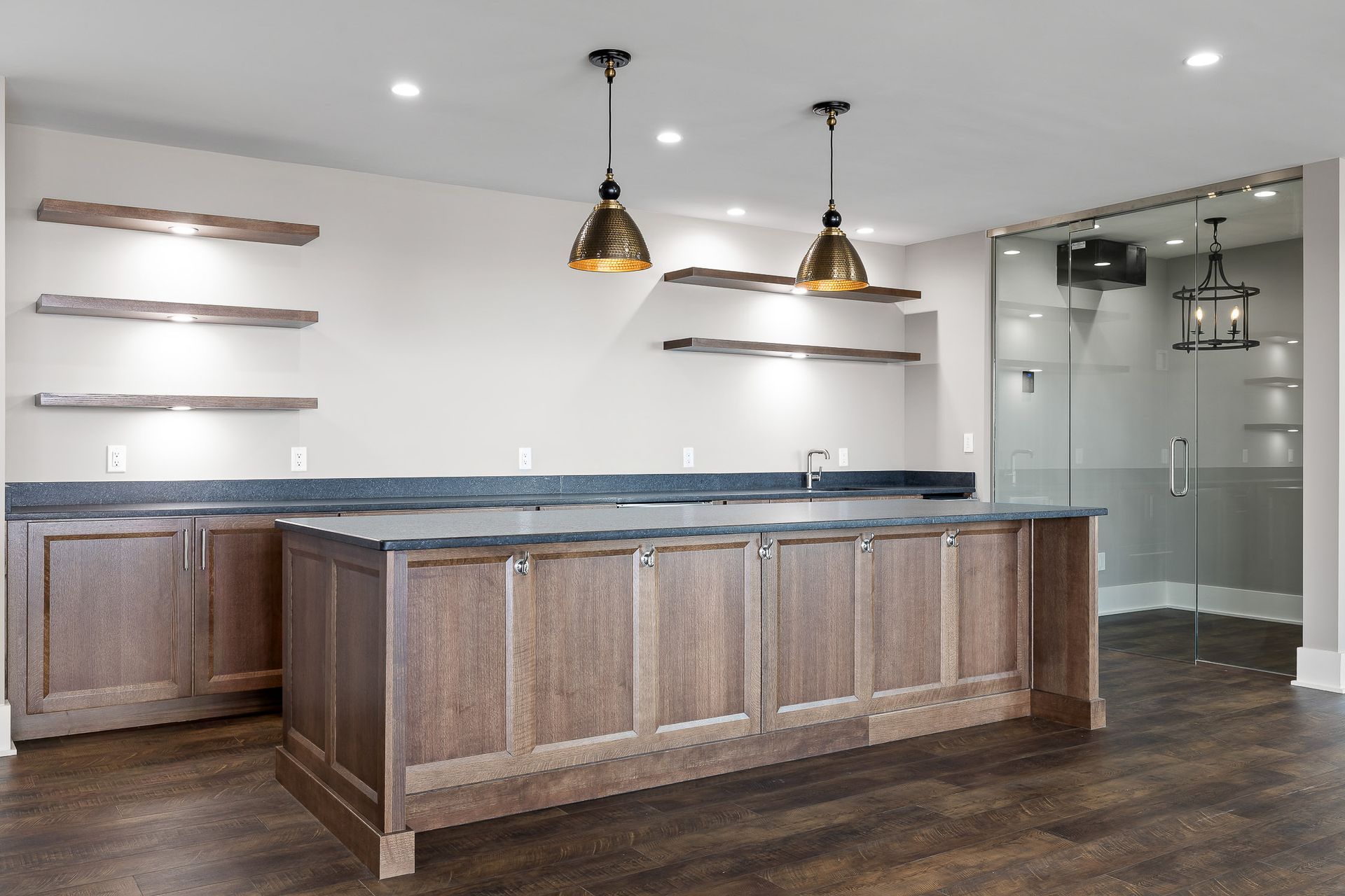 Basement bar with wooden cabinets, granite countertops, floating shelves, and pendant lights.
