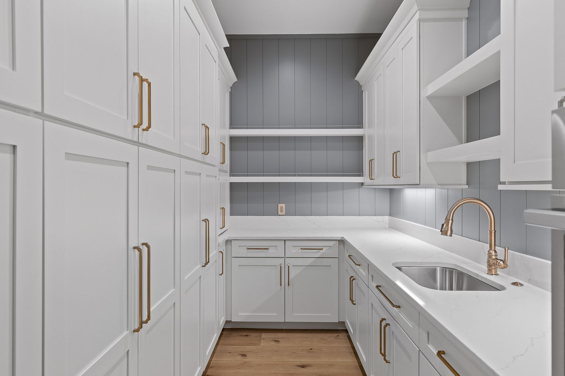 White pantry with cabinets, sink, shelves, and gold fixtures. Wooden floor.
