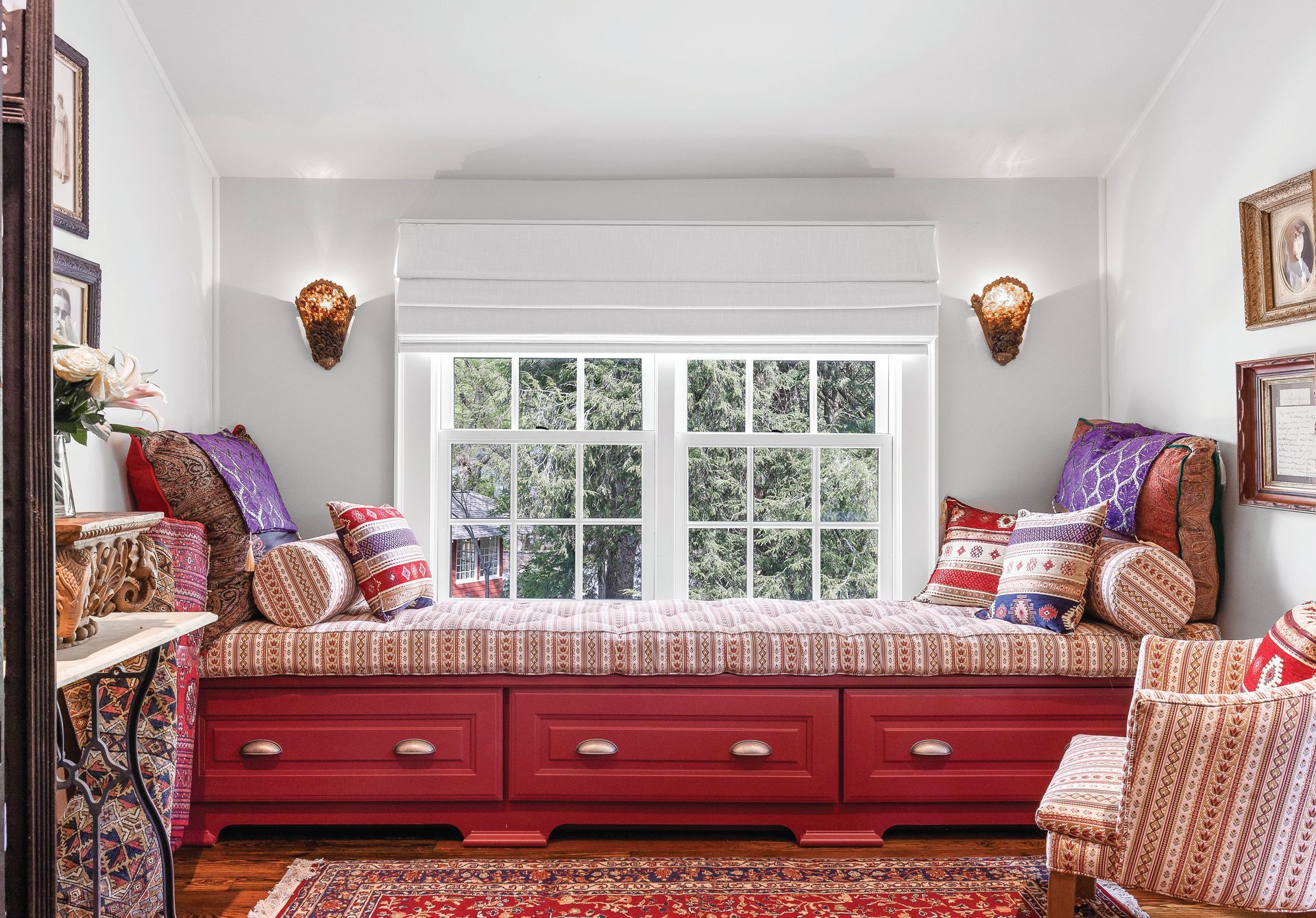 Cozy window seat with red storage drawers, patterned cushions, and decorative sconces; surrounded by artwork.