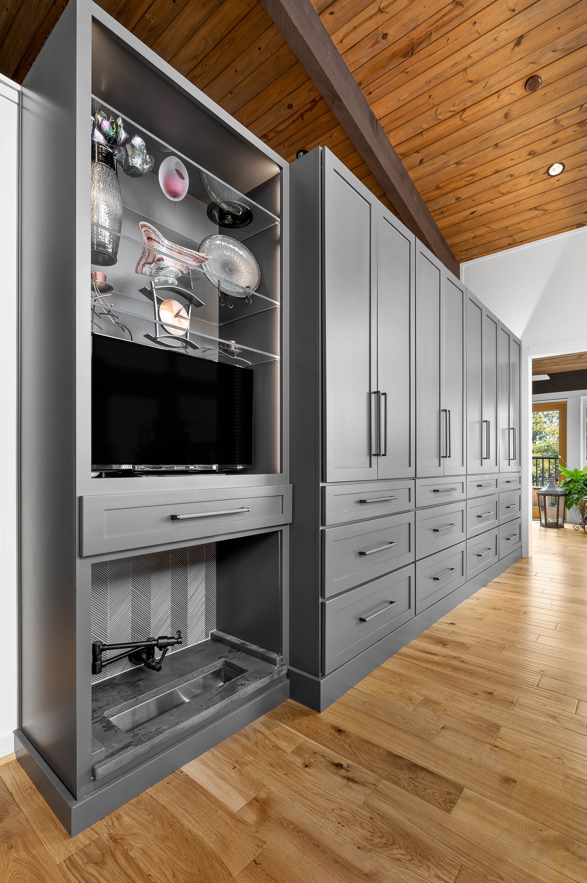 Gray built-in cabinets with a TV and glass shelving in a room with wooden floors and ceiling.
