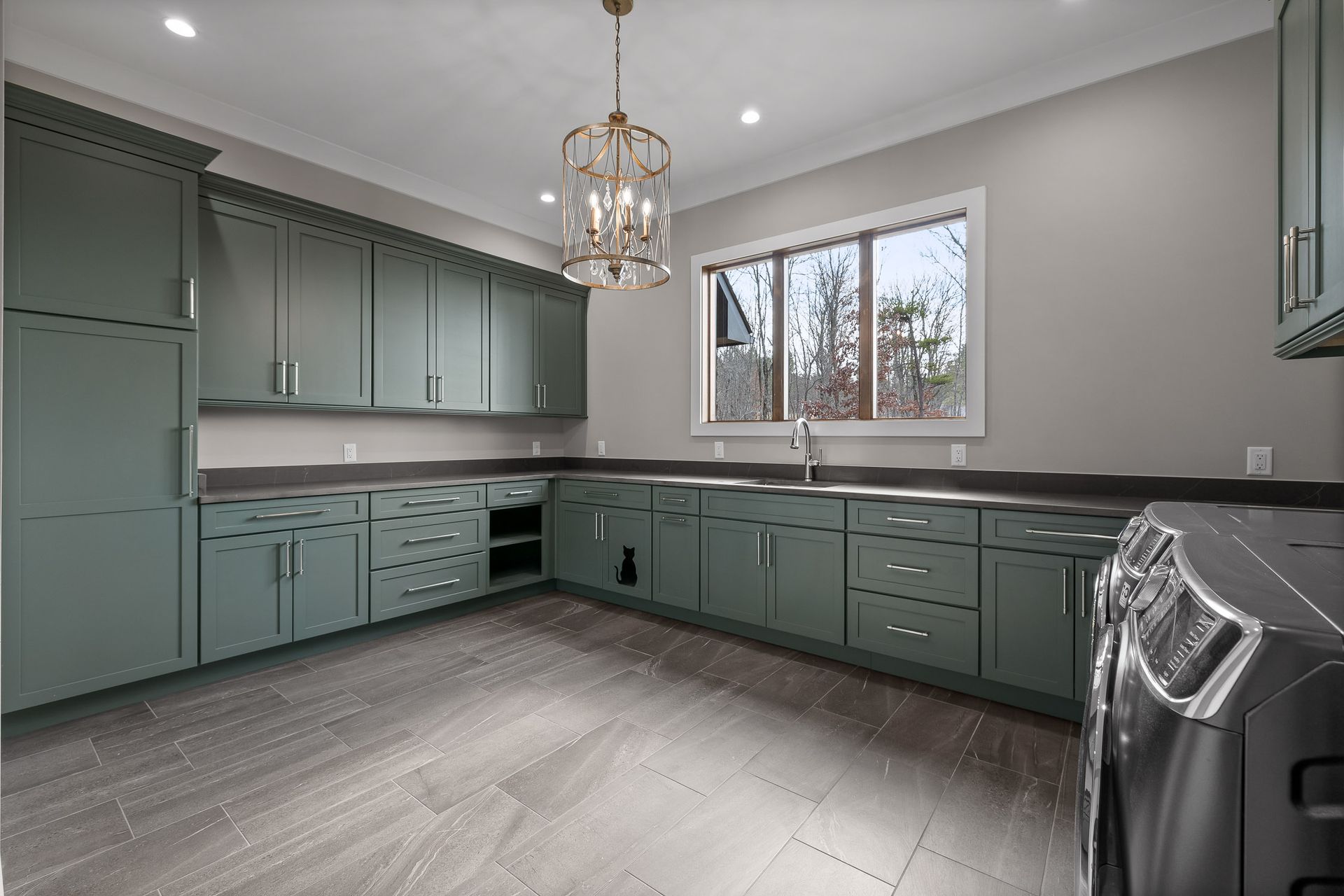 Spacious laundry room with green cabinets, gray countertops, gray floors, silver chandelier, and washer/dryer.