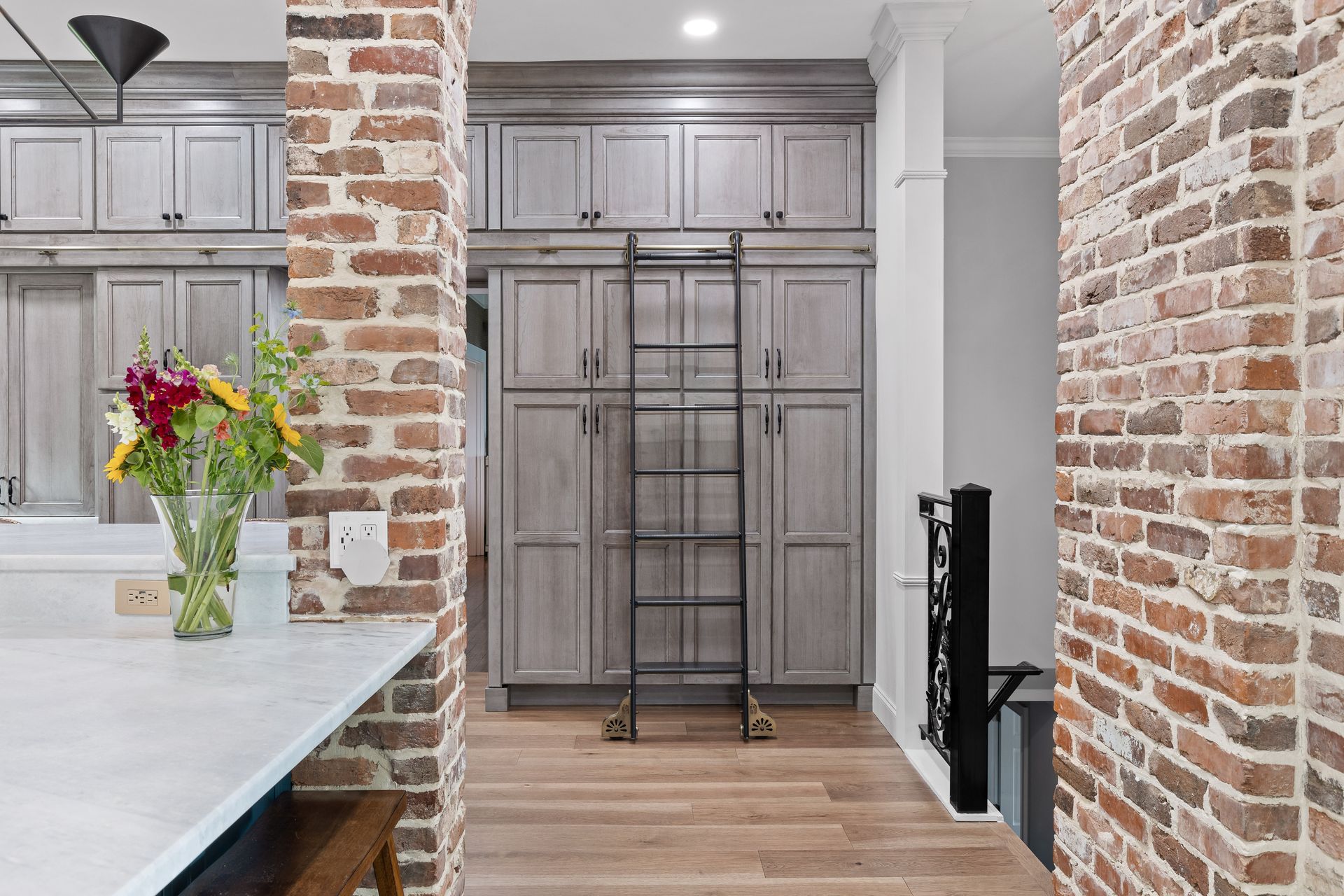 Kitchen with gray cabinets, exposed brick, ladder, and a vase of flowers.