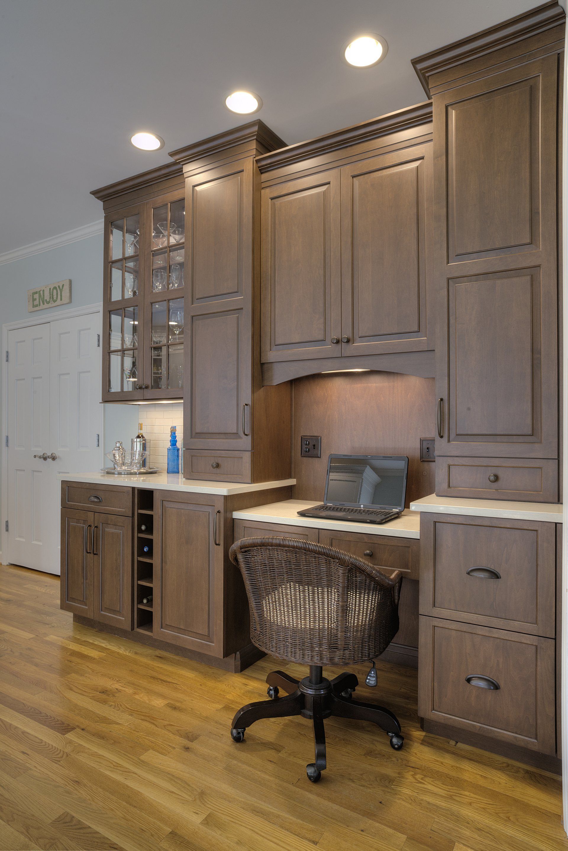 Home office nook with brown cabinets, laptop, and wicker chair on hardwood floor.