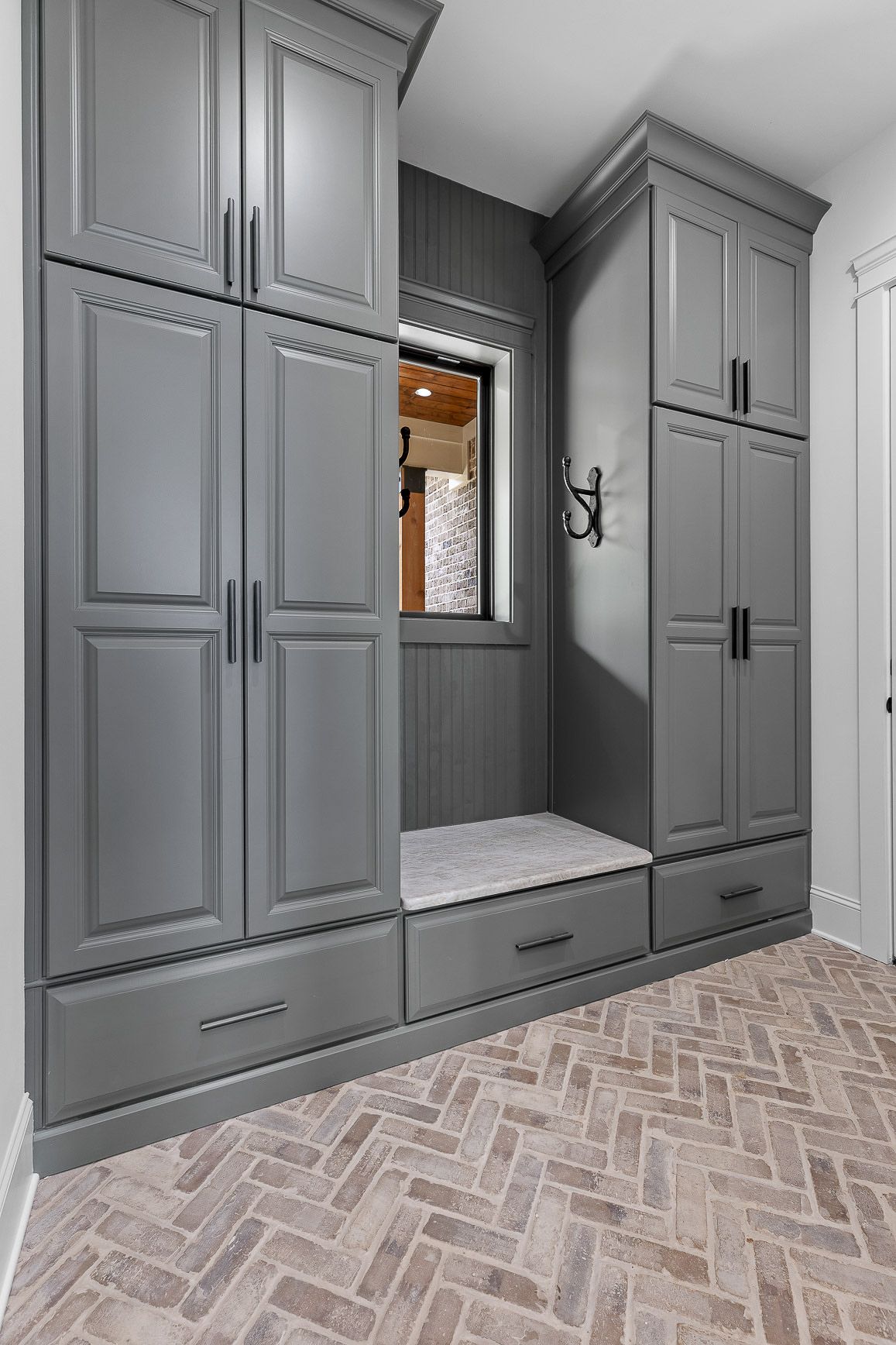Gray built-in storage with a window seat. Cabinets surround a bench on a herringbone brick floor.