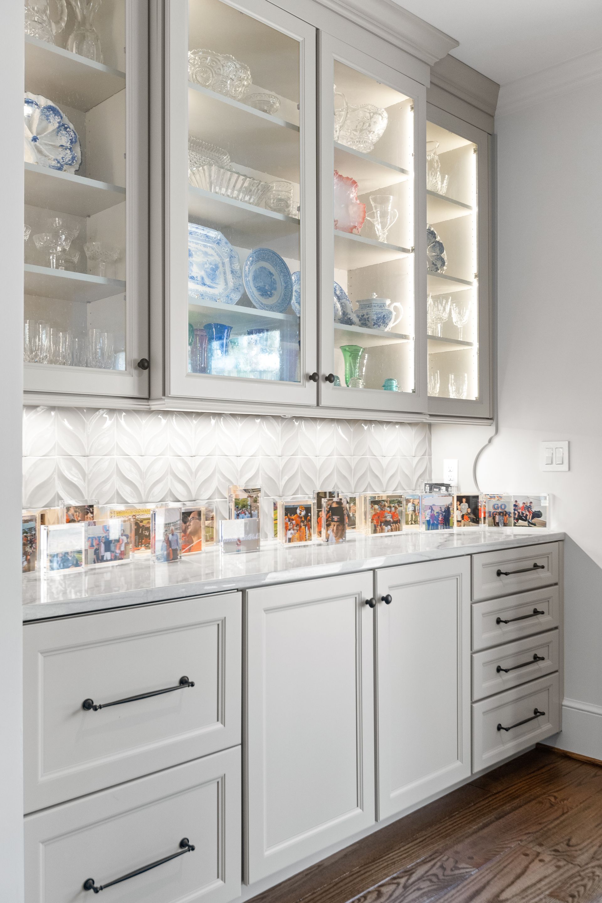Built-in white cabinets with glass-front upper cabinets displaying dishware, and a countertop with photos underneath.