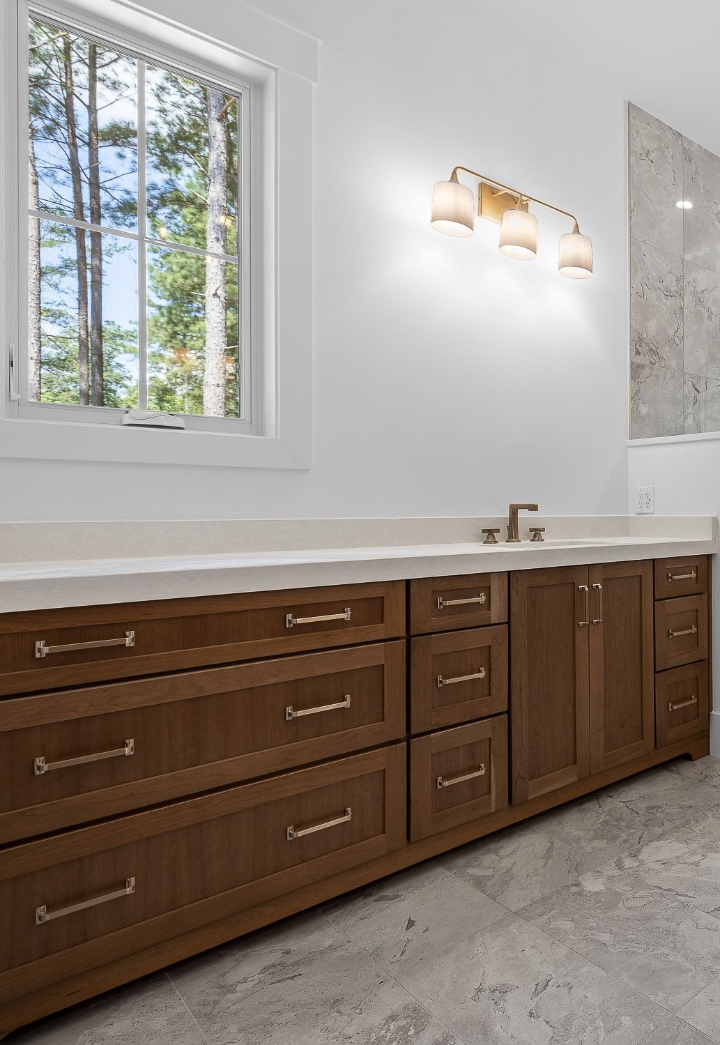 Bathroom with wood vanity, beige countertop, window, and gold light fixture.