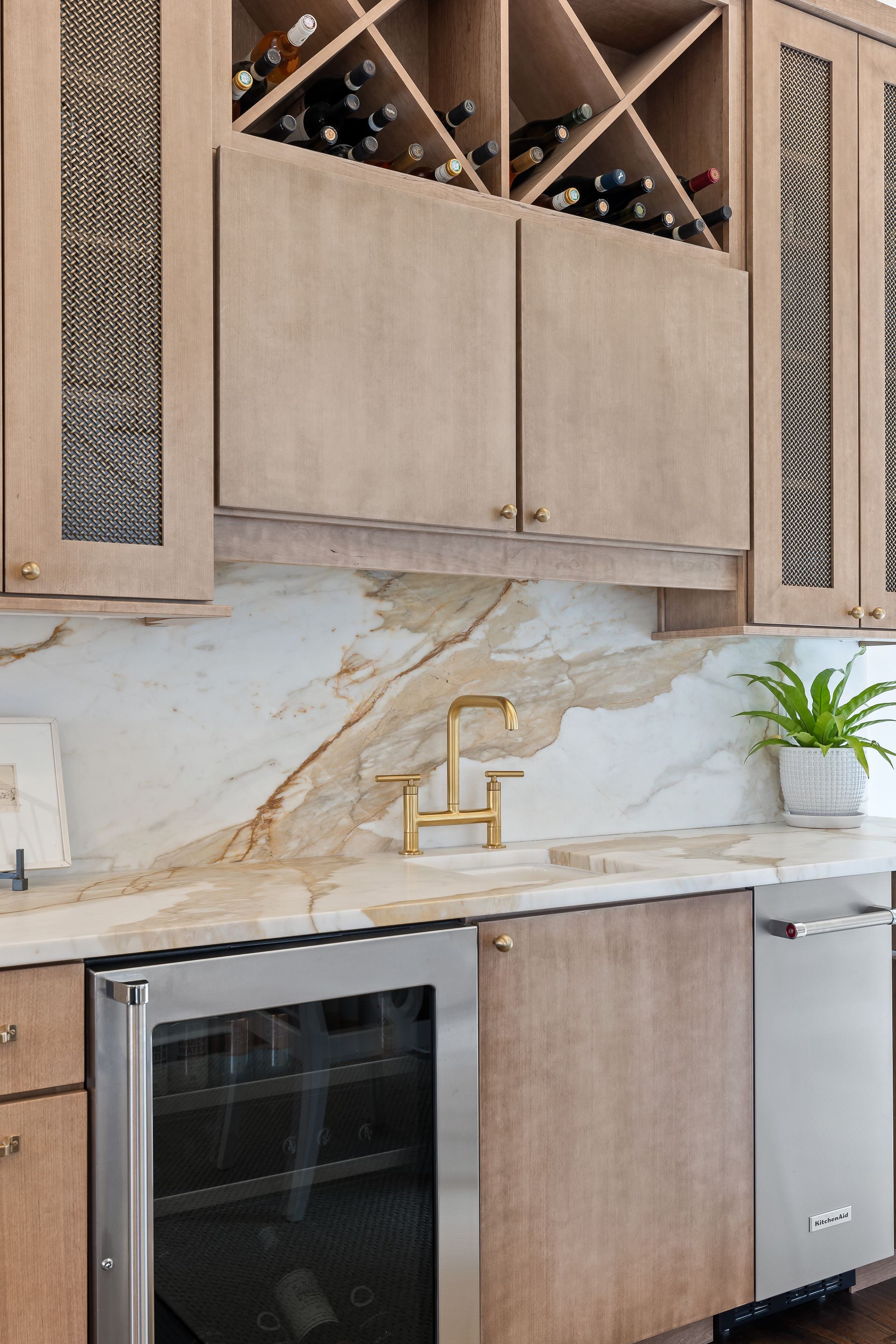 Wine bar with light wood cabinets, marble backsplash, wine fridge, and gold faucet.