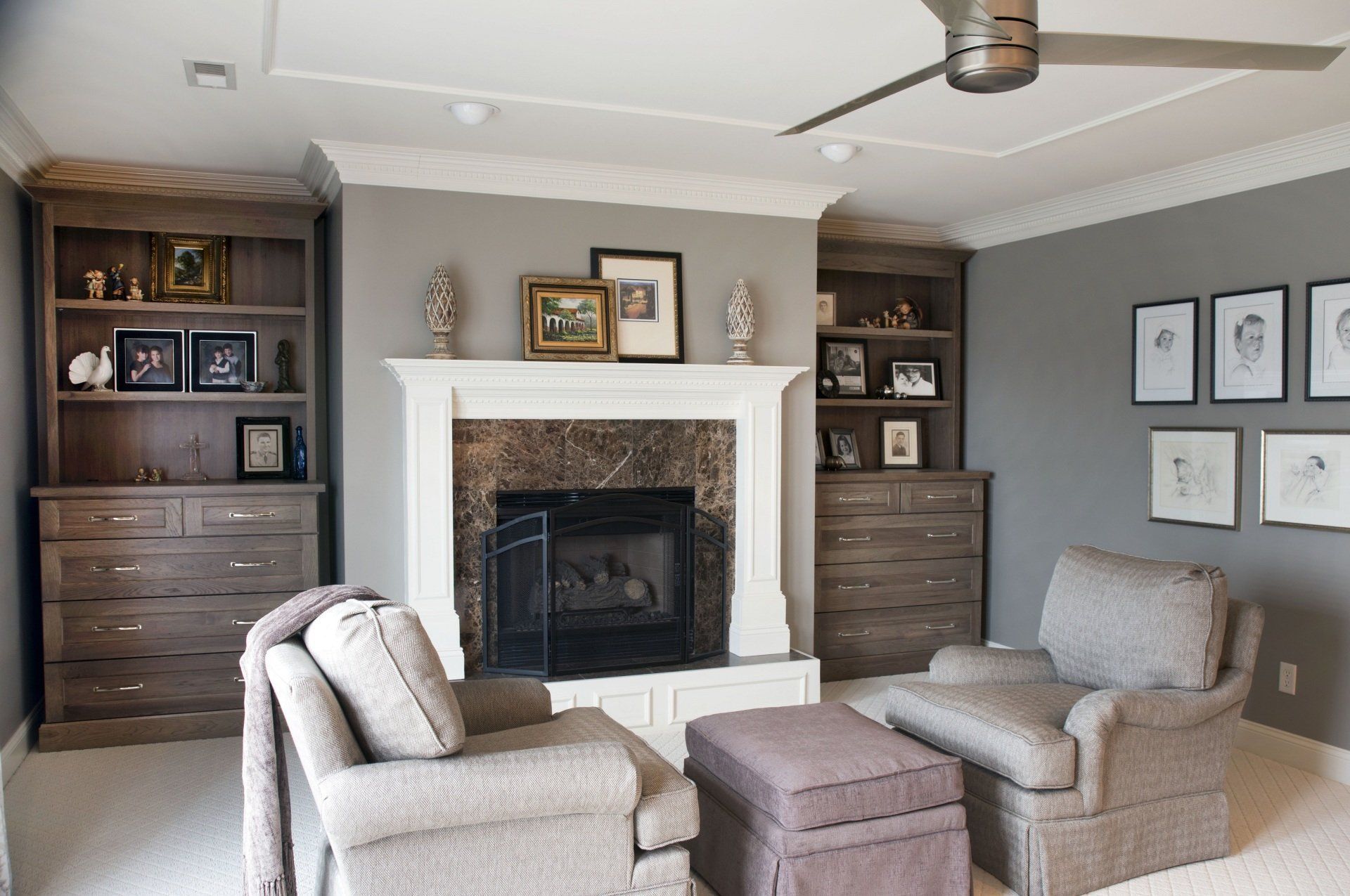 Cozy living room with a fireplace, built-in bookshelves, two armchairs, and an ottoman. Gray walls, light carpet.