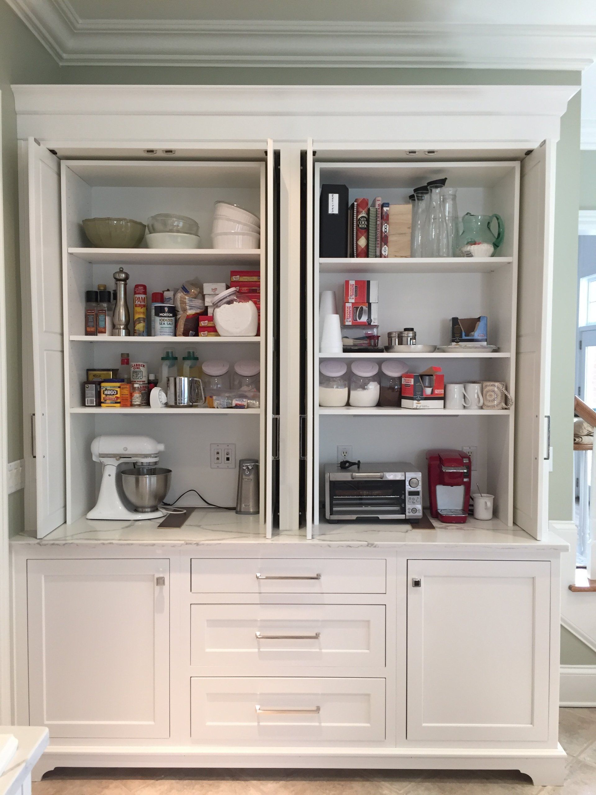 White kitchen pantry with pull-out shelves and countertop, filled with food, appliances and supplies.
