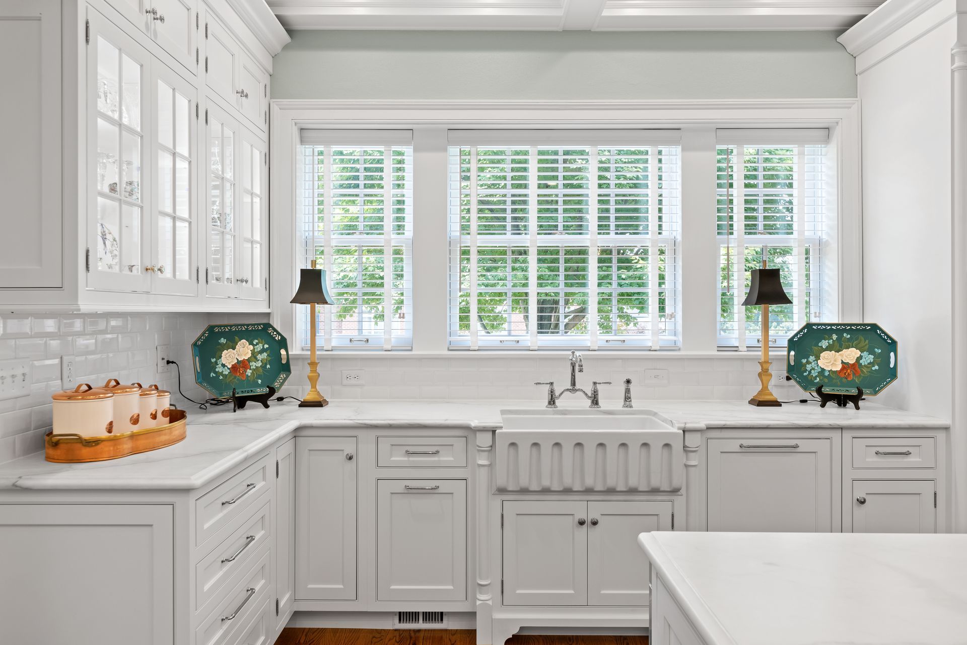 White kitchen with a farm sink, white cabinets, and three windows. Two lamps flank the windows.