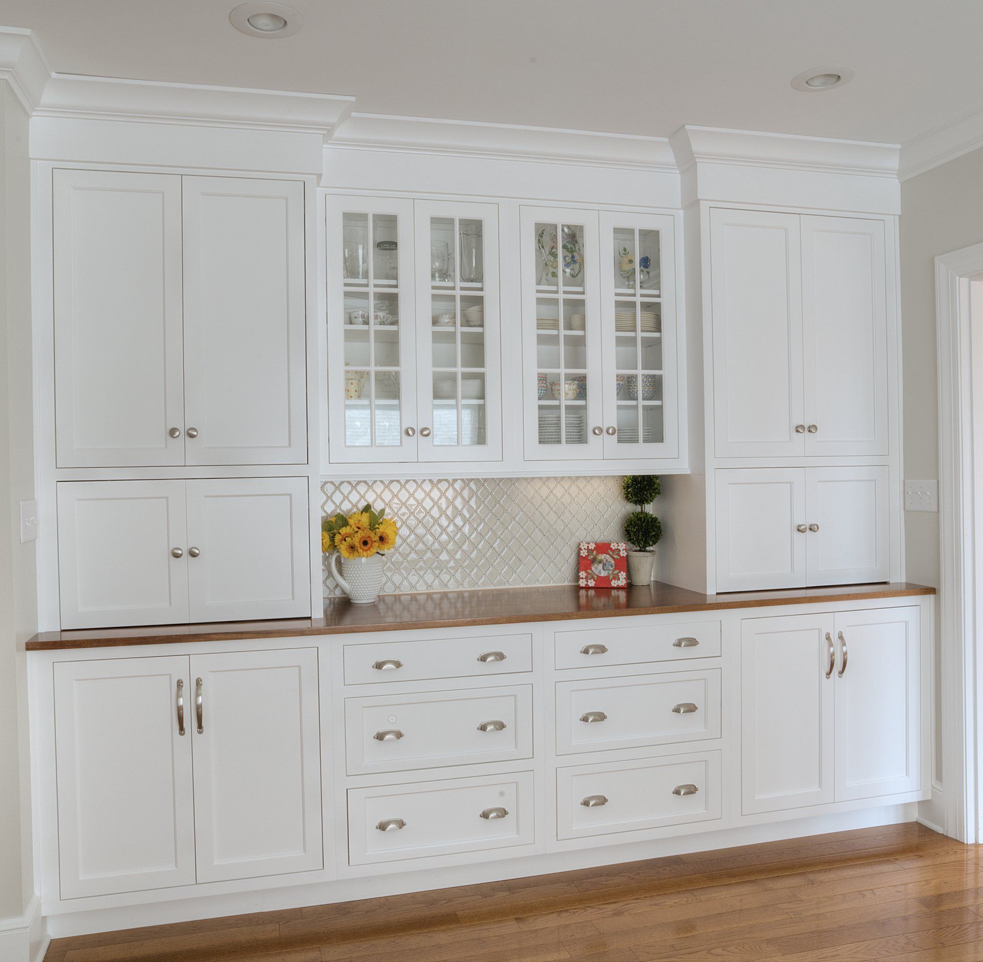 White kitchen cabinetry with glass-fronted uppers, drawers, and a wood countertop against a light-colored wall.