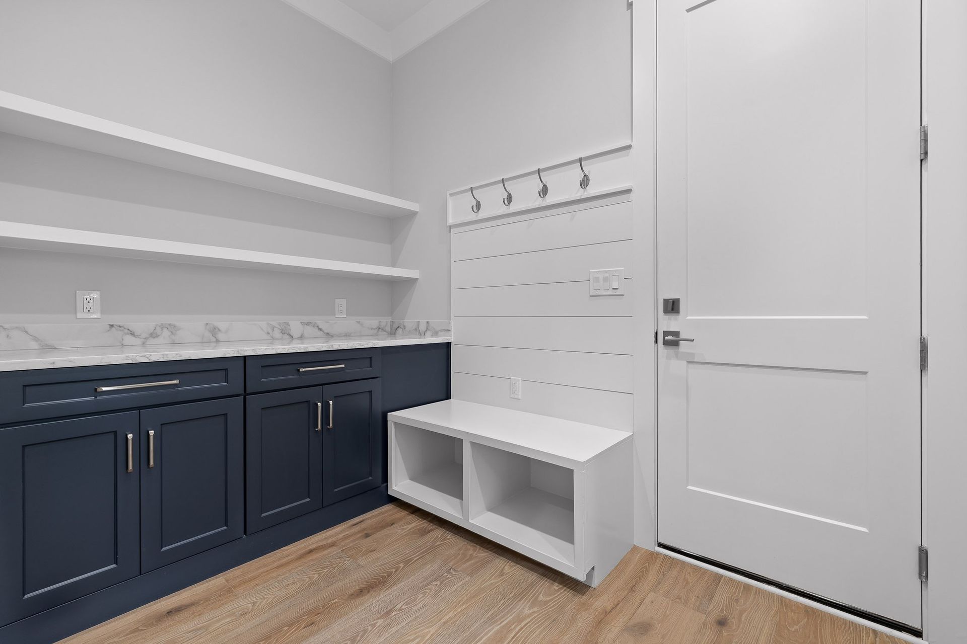 Mudroom with navy blue cabinets, white bench, and shelves; white door and light wood floor.