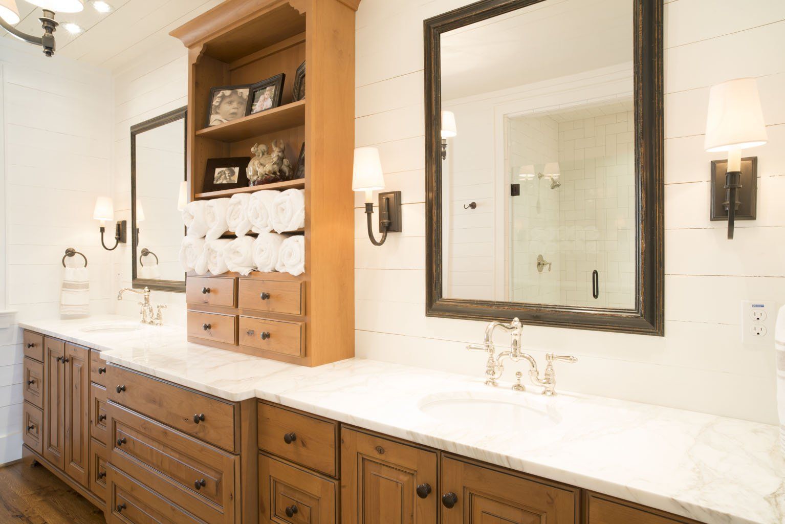 Bathroom with double sinks, wooden cabinets, marble countertop, two framed mirrors, and wooden bookcase.