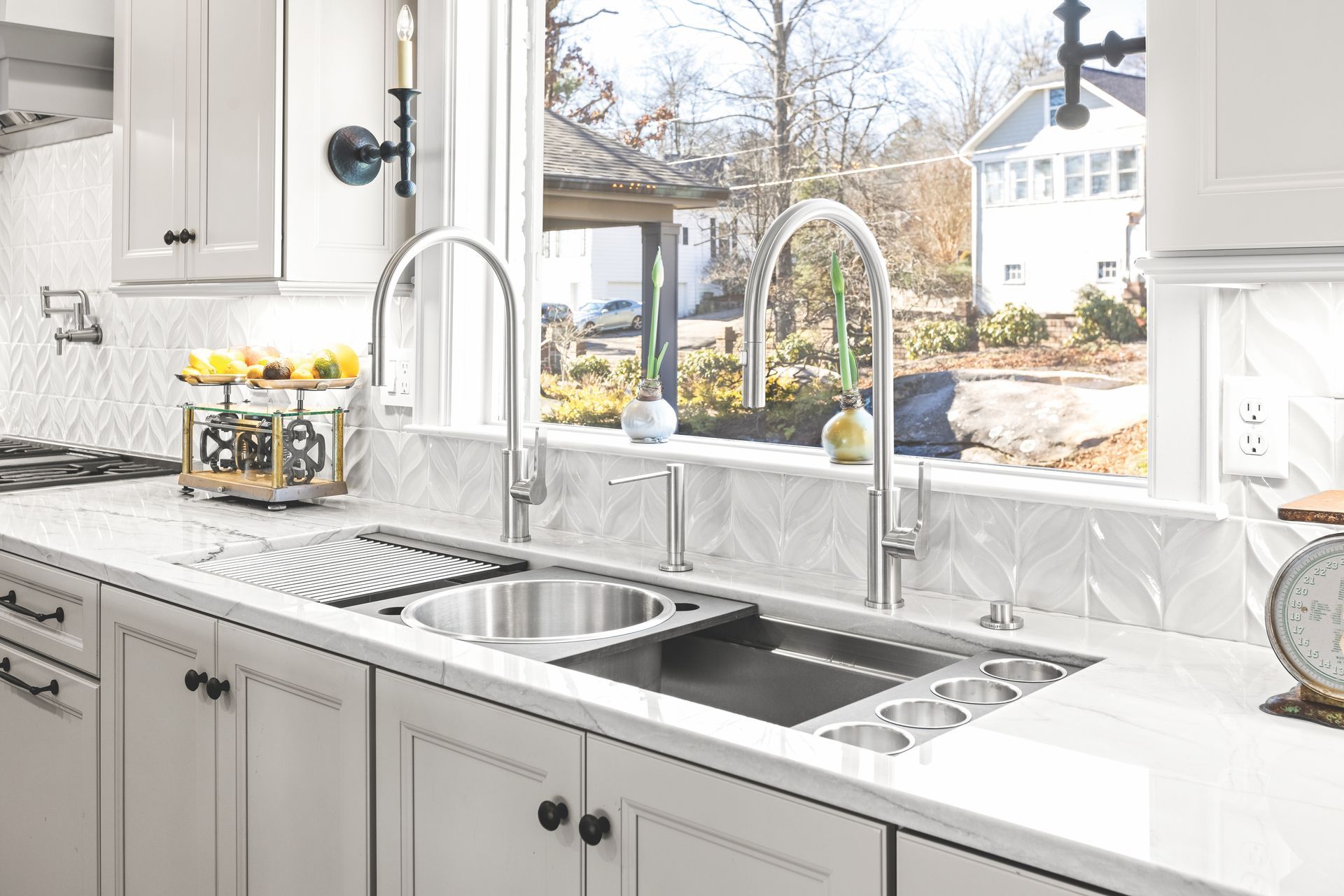 Kitchen sink with two faucets, looking out a window at houses, with white cabinets and countertops.