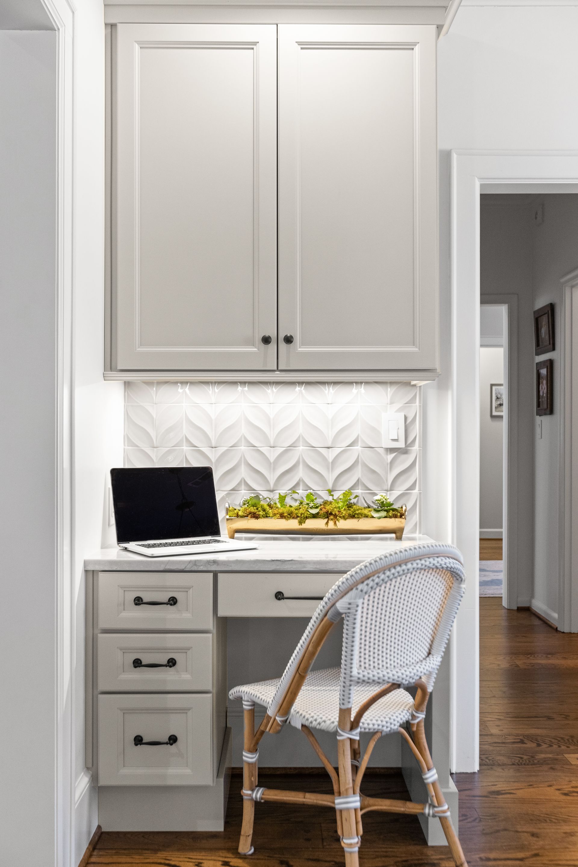 A small home office nook with a built-in desk and cabinets. Light gray and white color scheme, with a wicker chair.