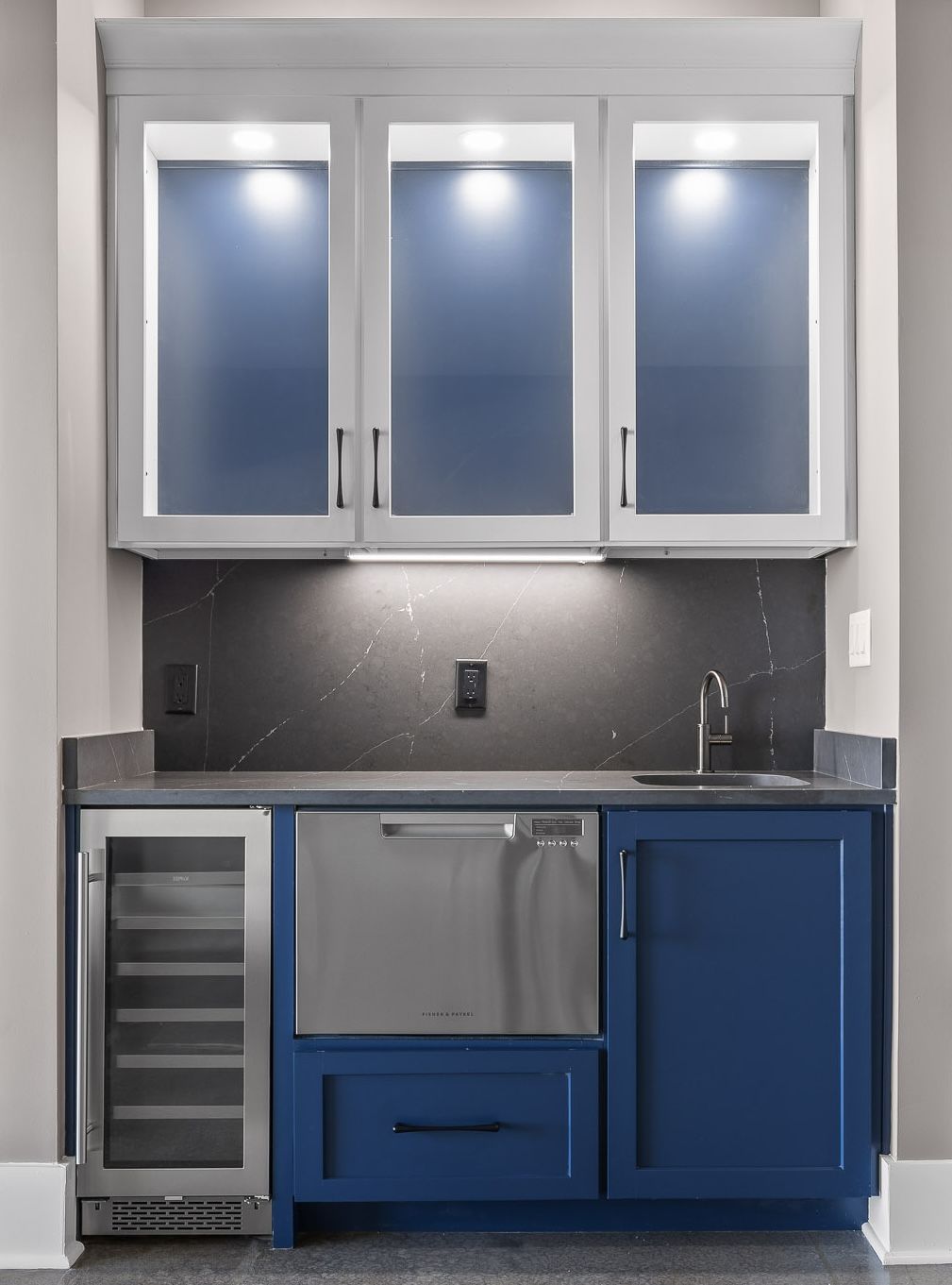Blue and gray bar with wine fridge, dishwasher, sink, and upper cabinets with lights.