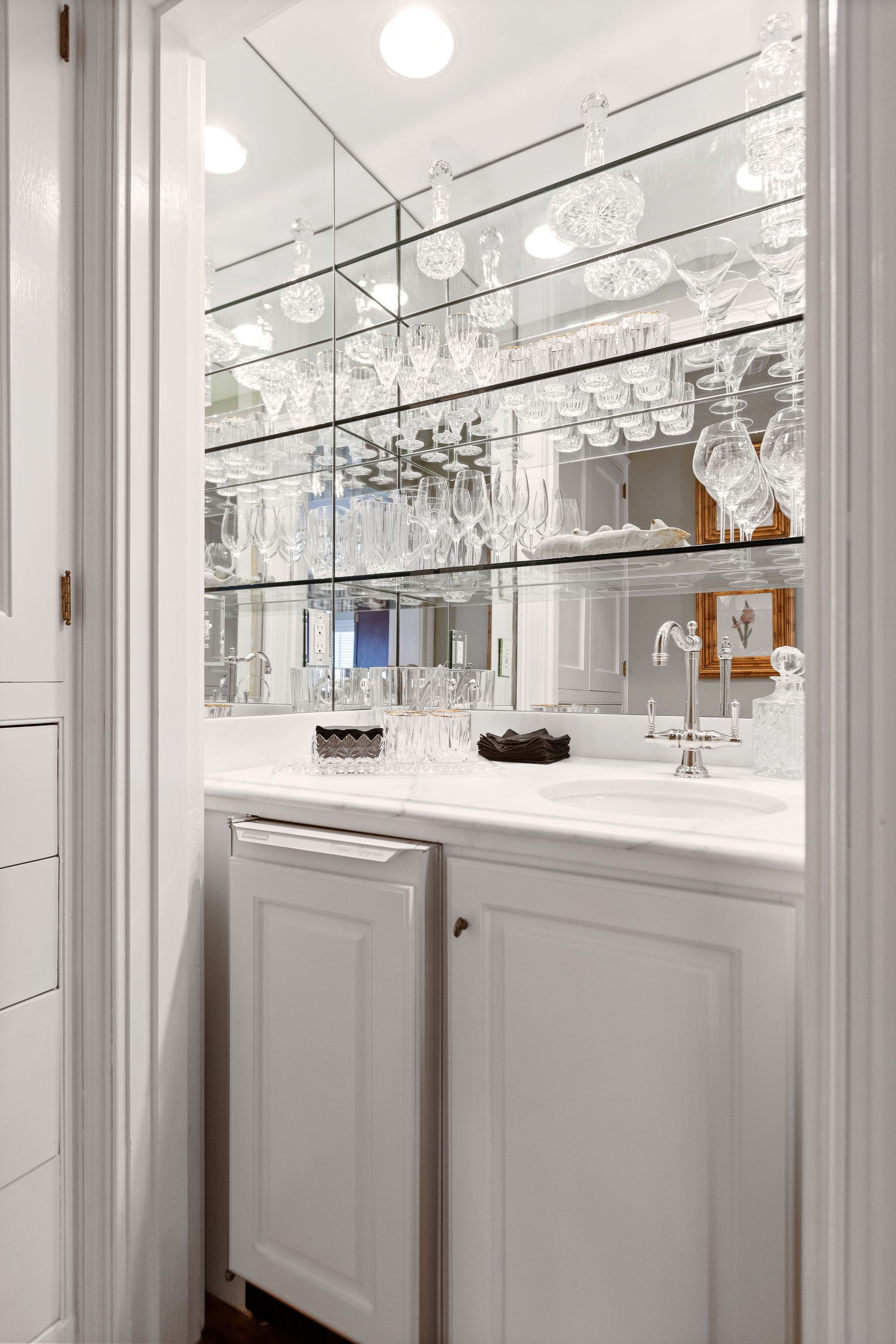 White-cabinet bar with a mirrored back wall, shelves of glassware, and a marble countertop.