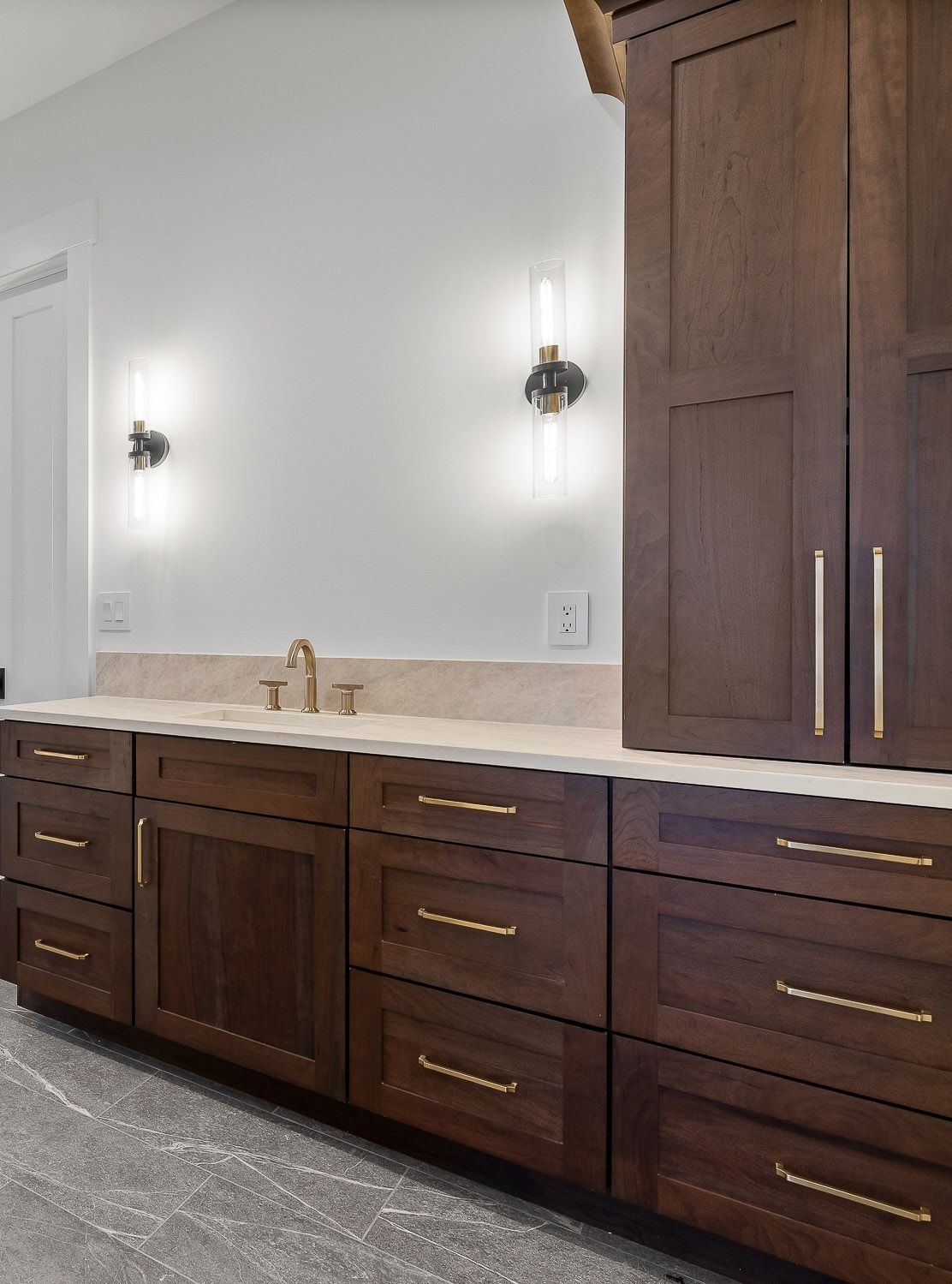 Dark wood bathroom vanity with gold fixtures, white countertop, and sconce lighting.